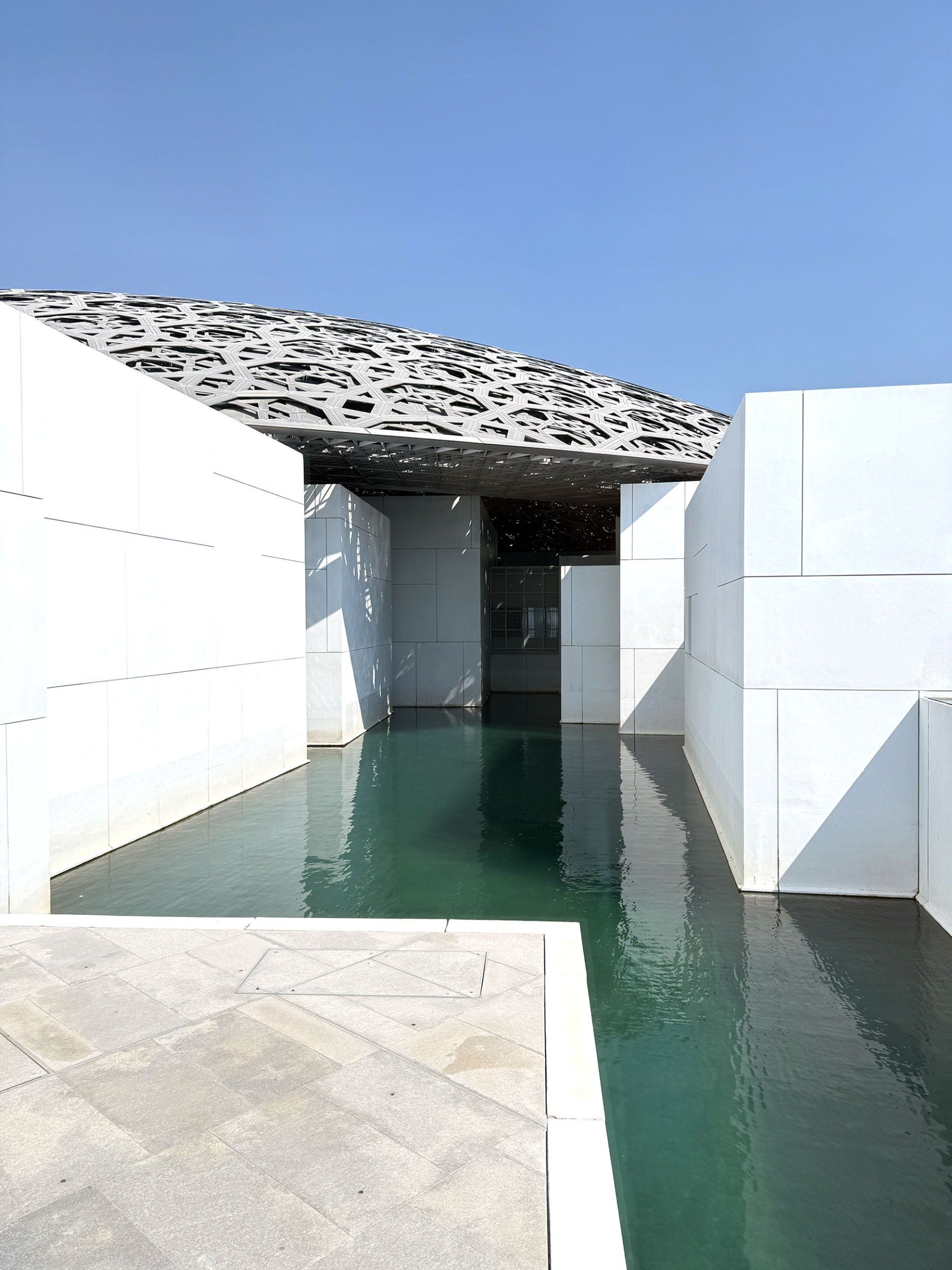 L'image montre une structure moderne et architecturale. À l'avant-plan, on voit une surface d'eau calme, reflétant le ciel bleu. De chaque côté de l'eau, il y a des murs blancs aux lignes épurées, créant un contraste avec le toit, qui est de forme organique et complété par des motifs découpés. Le design est minimaliste, mettant en valeur la lumière naturelle et l'harmonie avec l'environnement. L'ensemble dégage une ambiance de sérénité et de modernité.