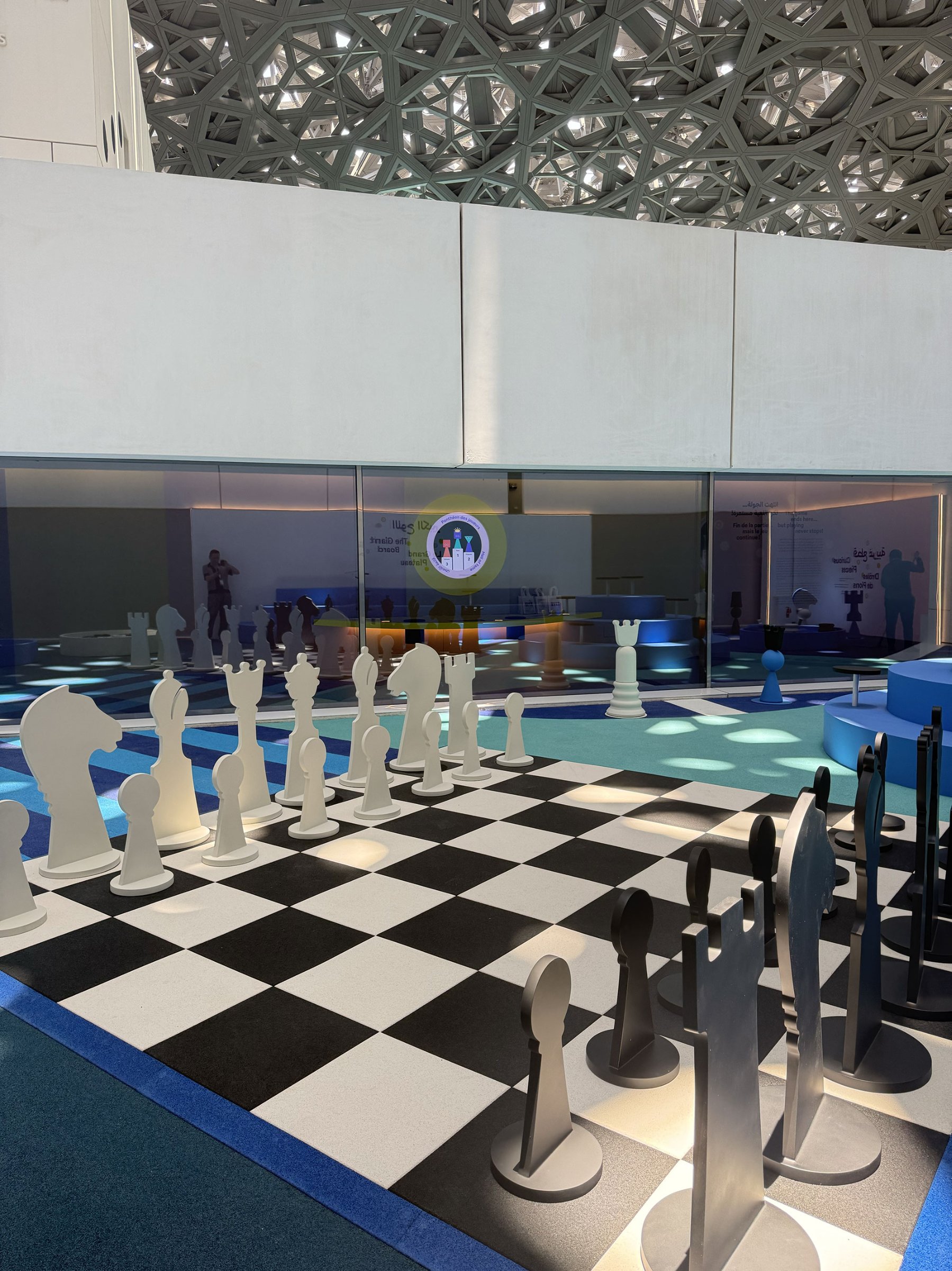 L'image montre un espace de jeu d'échecs intérieur. On y voit un grand plateau d'échecs au sol, avec des pièces grandeur nature en noir et en blanc. L'environnement est moderne et lumineux, avec des éléments architecturaux intéressants visibles au plafond. À l'arrière-plan, on aperçoit une zone de présentation ou d'information, avec des silhouettes de personnes. Le tout crée une atmosphère ludique et interactive.