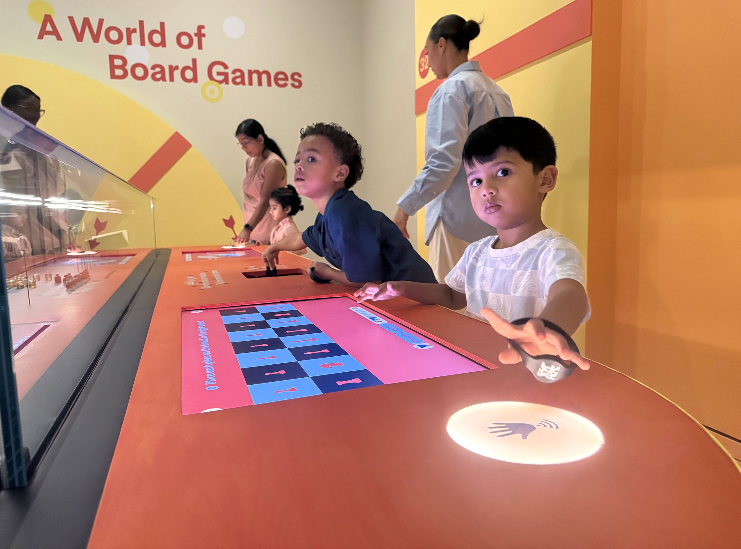 L'image montre un espace interactif dédié aux jeux de société. On y voit plusieurs enfants, dont deux garçons qui semblent très intéressés par les activités proposées. Ils touchent un écran tactile qui affiche des graphiques colorés. En arrière-plan, des adultes surveillent les enfants, et l'environnement est lumineux et accueillant, avec des murs aux couleurs vives. Les enfants interagissent avec des éléments numériques, suggérant un aspect éducatif et ludique de l'installation.