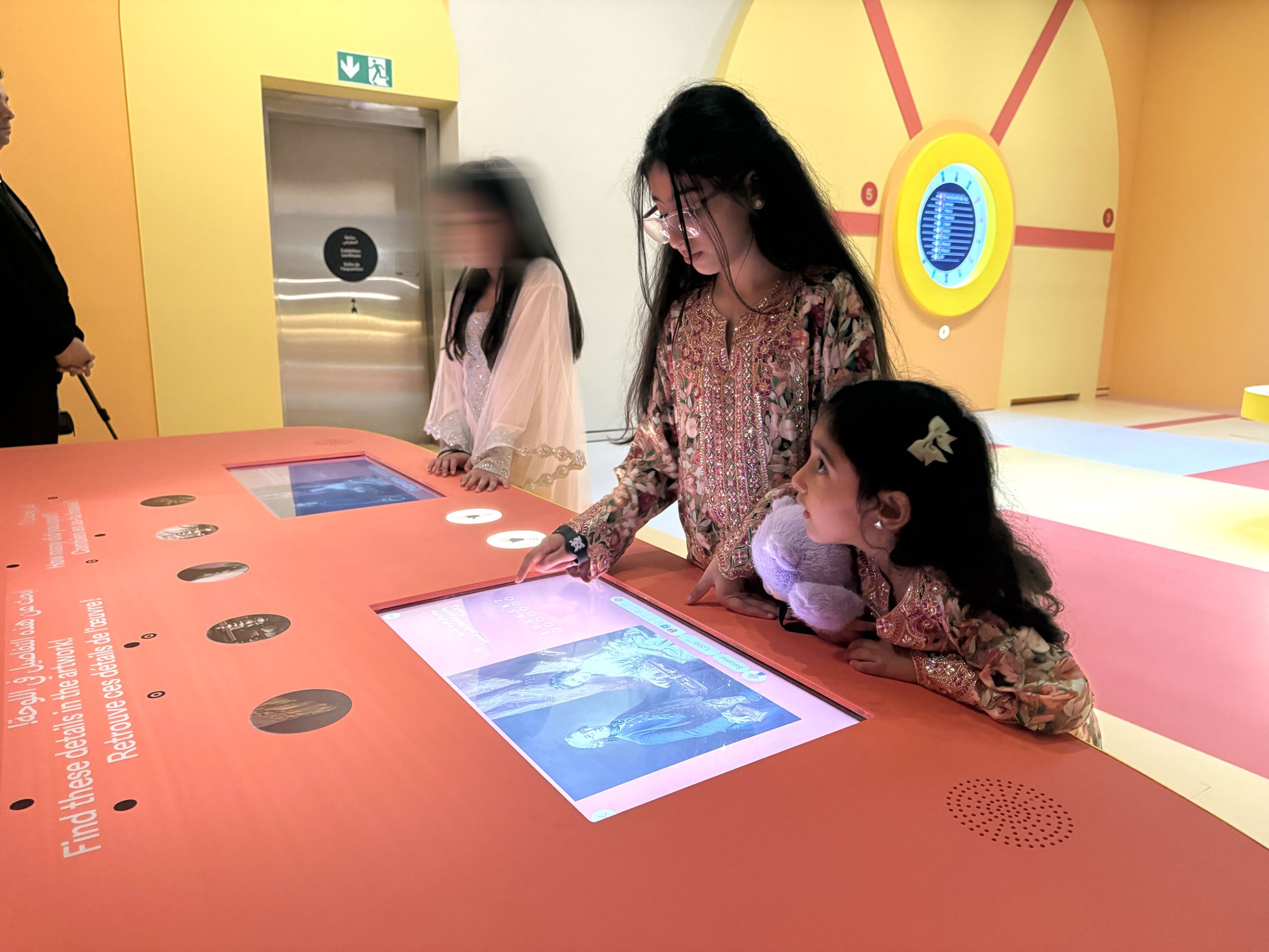 L'image montre un espace interactif où des enfants explorent une activité. Au centre, une jeune fille travaille sur un écran tactile, annotant ou interagissant avec des contenus numériques. À côté d'elle, une autre petite fille observe attentivement, tenant une peluche. En arrière-plan, on peut apercevoir d'autres personnes, mais leurs visages ne sont pas clairement visibles. L'environnement semble accueillant et coloré, suggérant qu'il s'agit d'une exposition ou d'un musée destiné aux enfants.