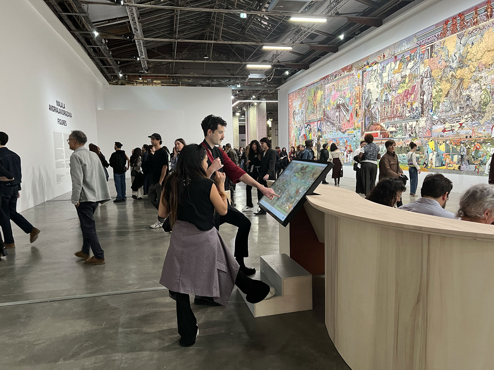 L'image montre une exposition artistique dans un grand espace. On peut voir un mur rempli de panneaux illustrés, avec une œuvre colorée qui attire l'attention. Au milieu, un groupe de personnes interagit avec un écran tactile, tandis que d'autres spectateurs explorent les environs. L'ambiance est animée, avec des visiteurs de tous âges qui découvrent les œuvres exposées. La lumière est vive et met en valeur les détails du décor.