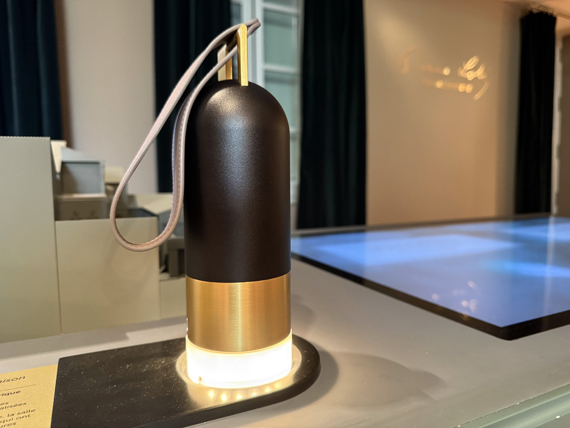 L'image montre un objet qui ressemble à une lampe ou un éclairage de table. Il a une forme cylindrique avec une partie supérieure en noir et une base en métal doré. Un cordon est attaché à la partie supérieure, ce qui suggère qu'il peut être déplacé facilement. La base est illuminée par une lumière douce, ajoutant une ambiance chaleureuse. L'objet est placé sur une surface moderne et épurée, avec un arrière-plan flou.