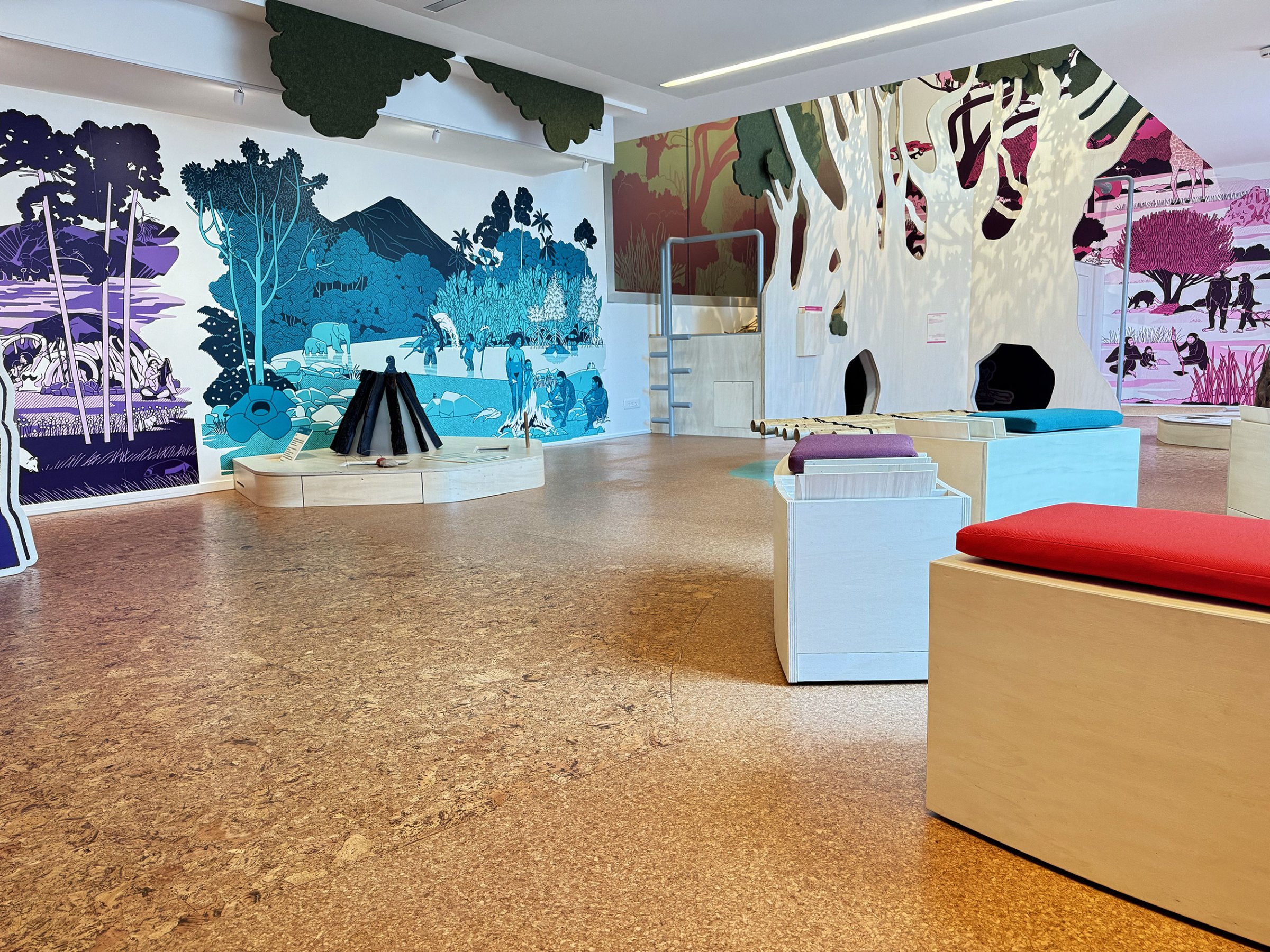 L'image montre un espace intérieur qui semble être une salle d'exposition ou un espace éducatif. Les murs sont décorés de grandes fresques colorées représentant des paysages et des scènes de nature, avec des arbres, des collines et des éléments animés. Au centre de la pièce, il y a plusieurs bancs en bois de différentes formes et couleurs, et une installation qui rappelle un feu de camp, posé au sol. L'éclairage est ample et met en valeur l'ambiance conviviale et accueillante de cet espace.