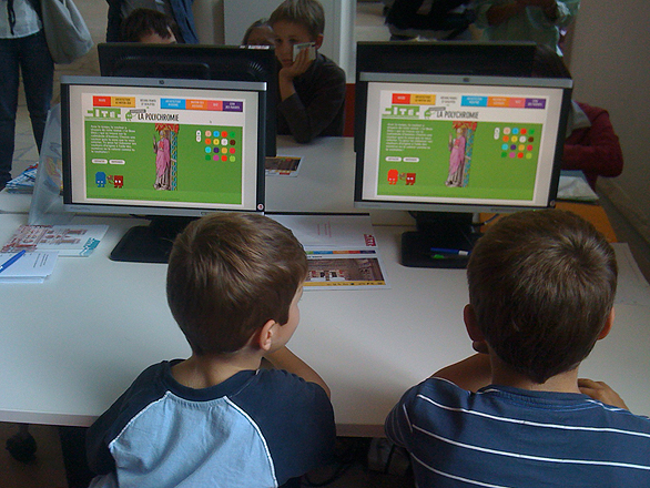 L'image montre un groupe d'enfants assis devant des ordinateurs, regardant l'écran. Les enfants sont concentrés sur une activité interactive qui semble être éducative, affichant des couleurs et peut-être des jeux. On aperçoit des affiches ou des ressources éducatives autour d'eux, suggérant un environnement d'apprentissage. Les enfants sont de dos, ce qui met l'accent sur leur engagement avec les ordinateurs.