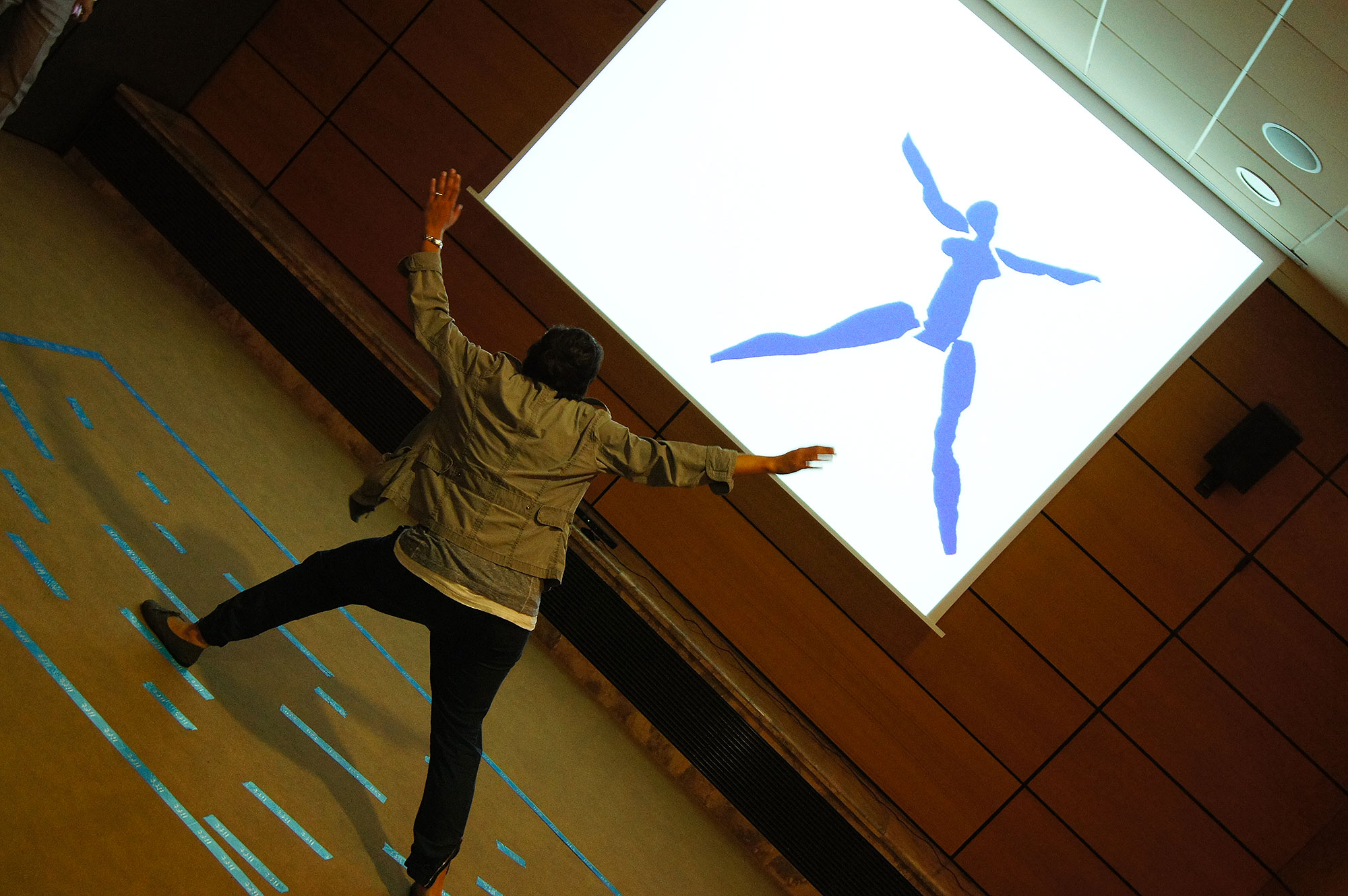 L'image montre une personne en mouvement, peut-être en train de danser ou de faire une performance, se tenant devant un grand écran. Sur cet écran, on voit une silhouette bleue représentant une figure humaine stylisée. L'environnement semble être une salle de réunion ou un espace de présentation, avec des murs en bois et un sol clair. La personne semble interagir avec l'image projetée, ajoutant une dynamique visuelle à la scène.