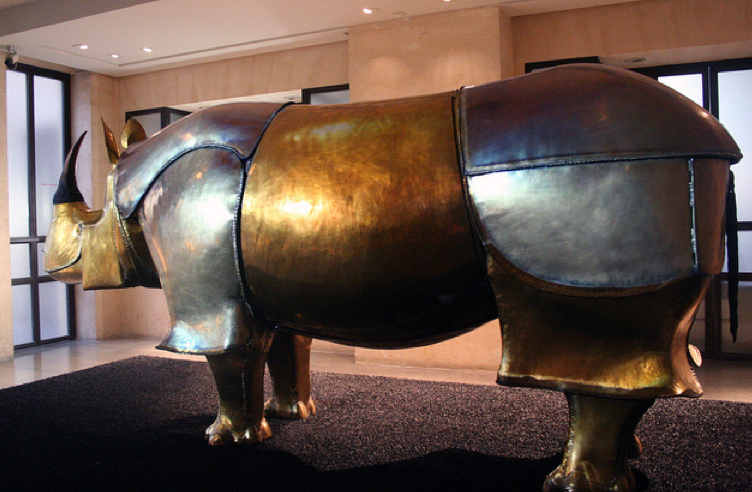 L'image montre une sculpture d'un rhinocéros, réalisée en métal poli. La sculpture a une finition dorée et argentée, ce qui lui donne un aspect brillant et artistique. Elle est placée dans un espace intérieur, probablement dans un musée ou une galerie d'art, avec un fond sobre et des lumières mettant en valeur les courbes de l'œuvre. Le rhinocéros se tient de profil, avec une posture imposante et élégante.