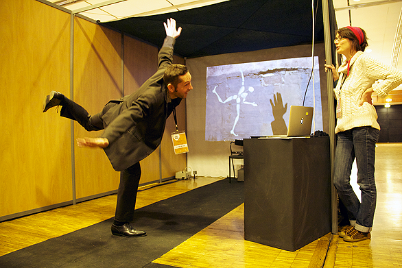 L'image montre un homme en costume qui effectue une pose de yoga ou d'équilibre, semblant s'appuyer sur une jambe tout en levant l'autre. Il est devant une table noire sur laquelle repose un ordinateur portable. À côté de lui, une femme avec des lunettes et un chapeau observe la scène. En arrière-plan, il y a une projection d'une image sur un mur, représentant des silhouettes ou des mouvements. L'ambiance semble créative et ludique.