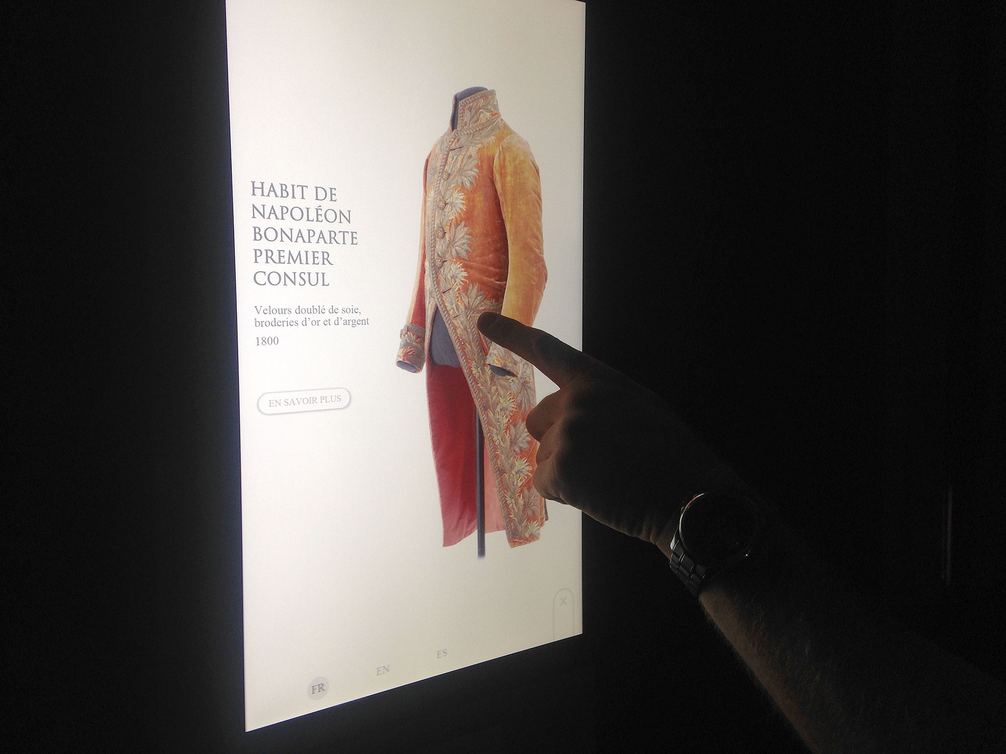 L'image montre un écran interactif présentant un habit de Napoléon Bonaparte, qui était Premier Consul. Le vêtement est richement décoré, en soie, avec des broderies. Une main se tient près de l'écran, soulignant le costume, et il y a une option pour en savoir plus en bas de l'image.