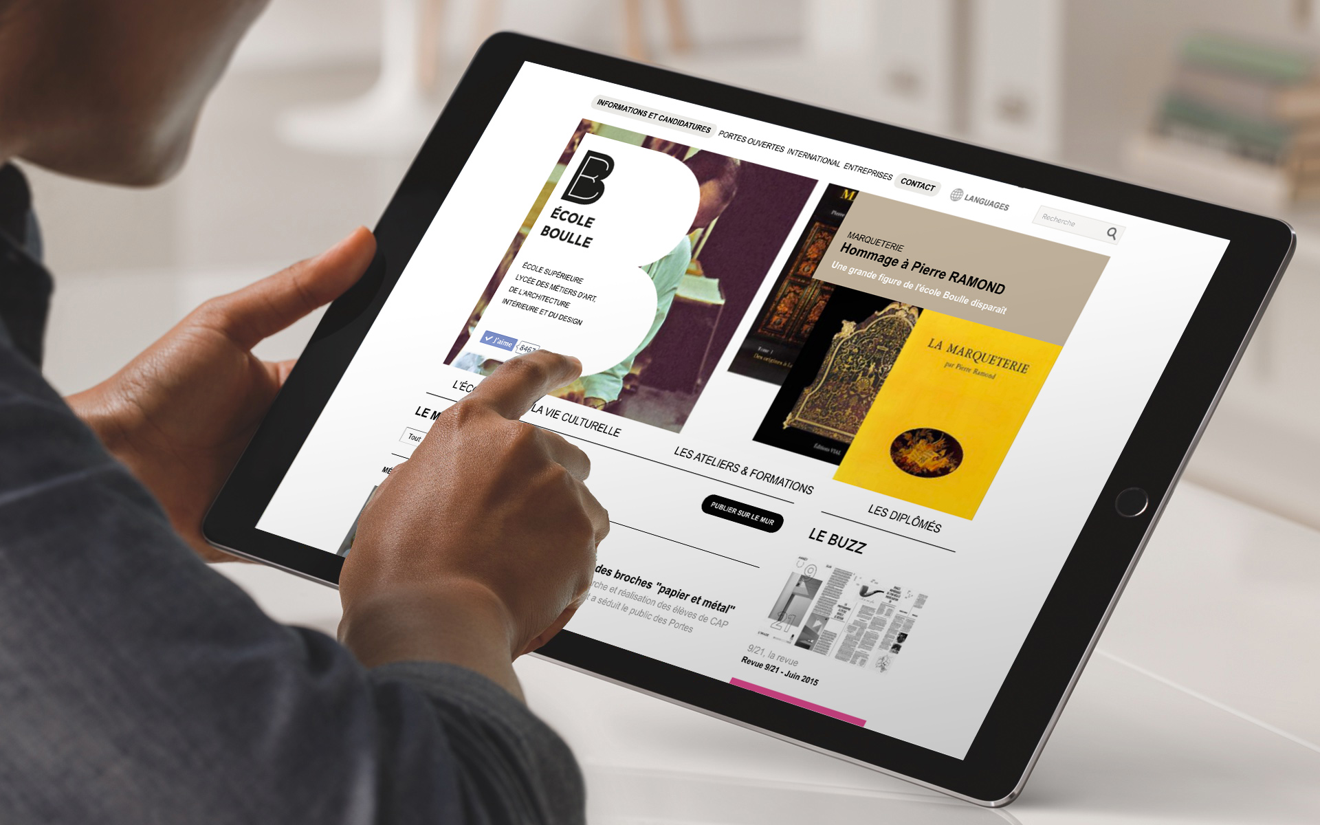 L'image montre une personne utilisant une tablette pour naviguer sur un site web. L'écran présente des informations sur une école, incluant des publications et des événements. On peut voir des visuels de livres et des sections comme "les expositions" et "le buzz". L'utilisateur semble intéressé par le contenu, interagissant avec l'écran tactile.