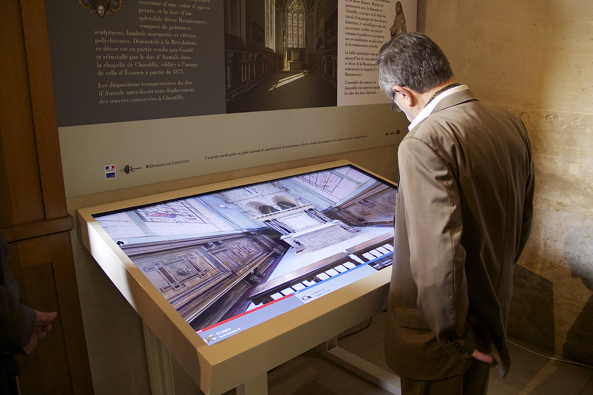 L'image montre un homme debout devant un grand écran tactile dans un espace intérieur. L'écran affiche une vue en 3D ou une reconstitution numérique d'une salle, probablement d'une église ou d'un lieu historique, mettant en évidence une architecture détaillée. L'environnement autour de l'écran est décoré de manière sobre, avec des murs en pierre et des éléments d'information visibles. L'homme semble attentif et intéressé par ce qu'il découvre à l'écran.