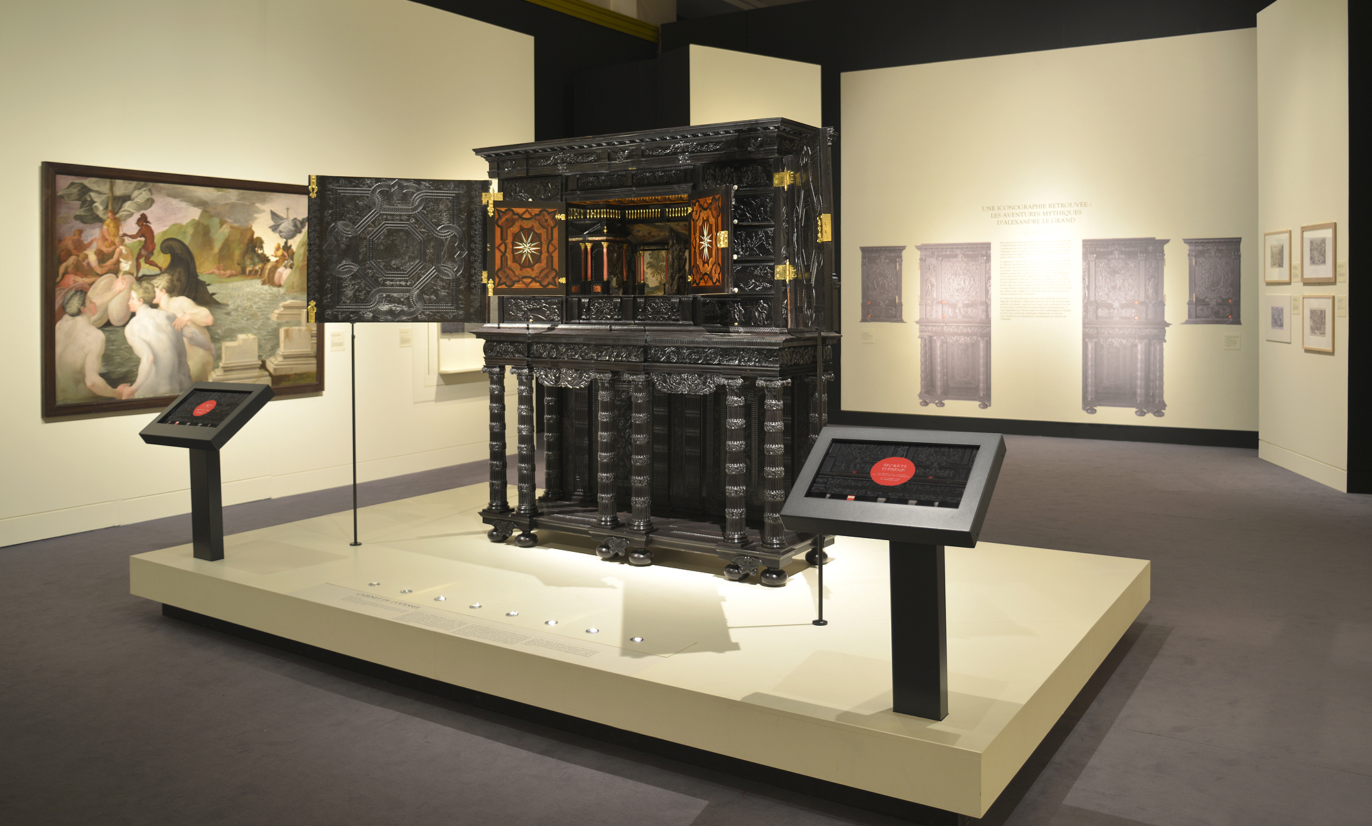 L'image montre une exposition muséale où un meuble ou une armoire ornée est mise en valeur au centre. Ce meuble, riche en détails, possède des sculptures et des marqueteries colorées. Des panneaux d'information sont positionnés près de l'objet, expliquant probablement son histoire ou son origine. À l'arrière-plan, on aperçoit un tableau dans un style classique, ainsi que des reproductions ou des œuvres similaires sur les murs. L'éclairage de l'espace met en avant les détails du meuble, créant une atmosphère d'admiration pour cet objet d'art.