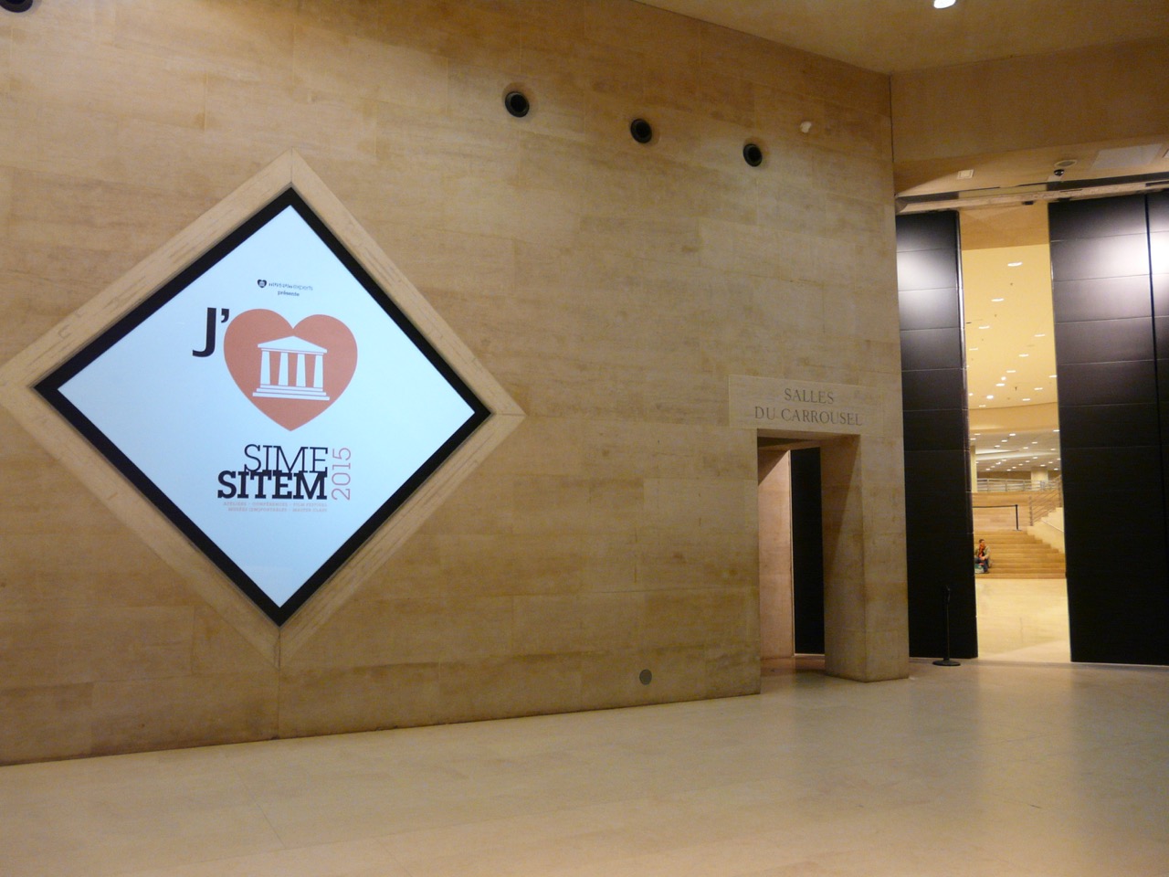 L'image montre un intérieur d'un bâtiment moderne, avec des murs en pierre claire. Sur le mur de gauche, il y a un grand écran affichant un logo avec un cœur et le texte "J' ♥ SITEM 2015". À droite, on aperçoit une entrée marquée "SALLES DU CARROUSEL", qui semble mener à un espace supplémentaire. L'éclairage est doux et crée une ambiance accueillante.