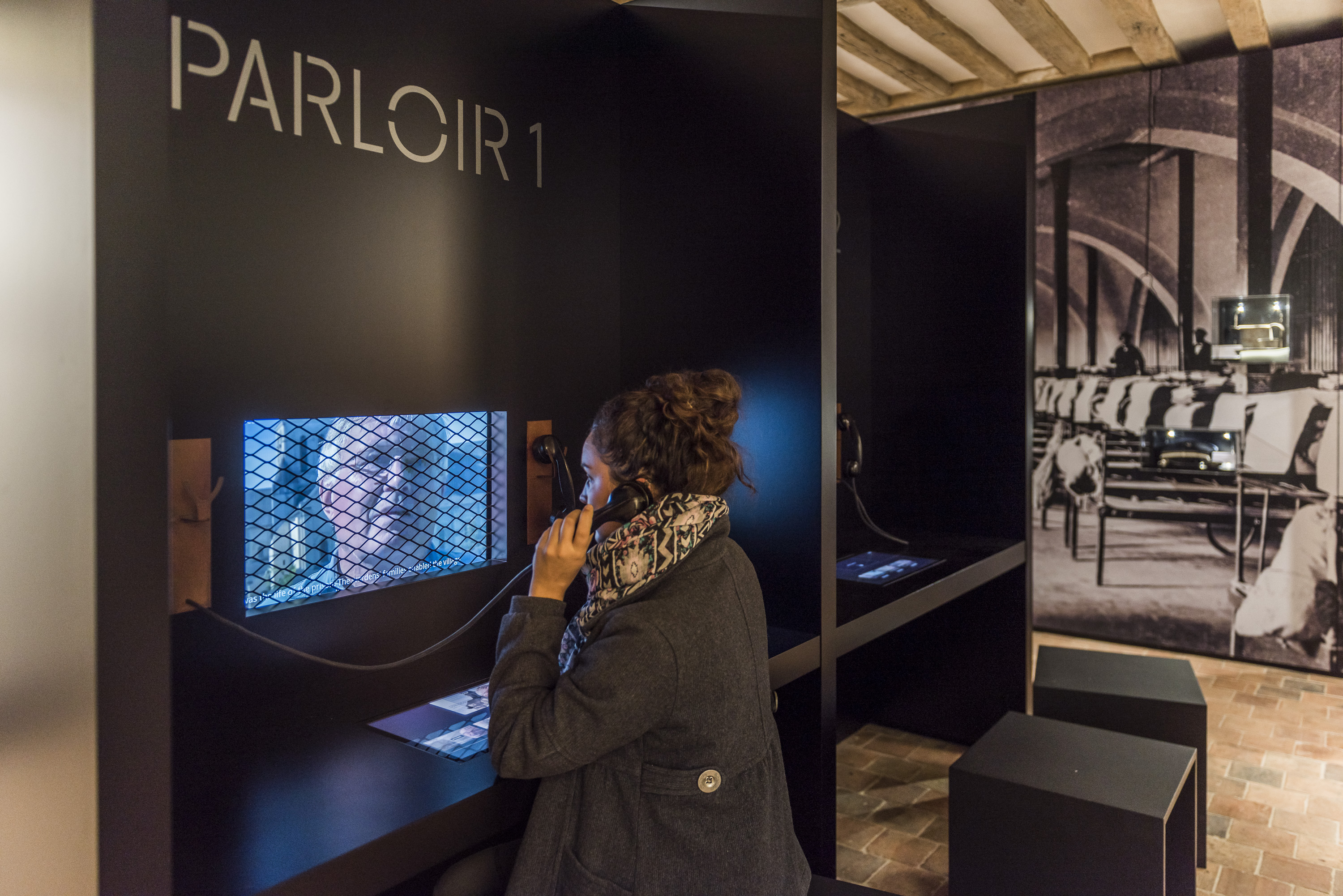 L'image montre un espace intérieur avec des cabines ou des sections séparées par des panneaux noirs. Une femme est assise à une table, utilisant un téléphone. Elle porte un manteau et une écharpe. À l'arrière-plan, on aperçoit des chaises alignées dans une pièce qui semble être un ancien lieu public ou historique. Un écran est visible, montrant une image à travers une grille. L'ambiance générale suggère un lieu de dialogue ou de communication, peut-être dans un cadre muséal ou éducatif.