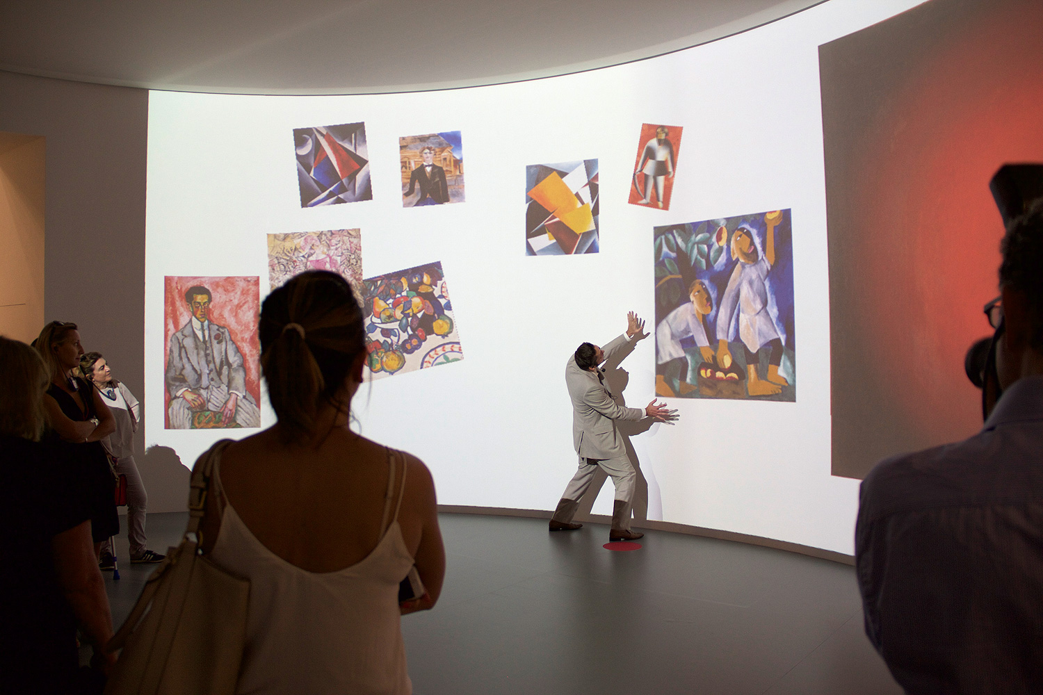 L'image montre une exposition artistique où un présentateur, habillé en costume clair, interagit avec des œuvres d'art projetées sur un mur courbe. Il semble expliquer ou mettre en valeur les installations, tandis qu'un groupe de visiteurs l'observe attentivement. Les œuvres affichées comprennent divers styles, tels que des peintures colorées et abstraites. L'ambiance est dynamique et immersive, illustrant une rencontre entre l'art et le public.