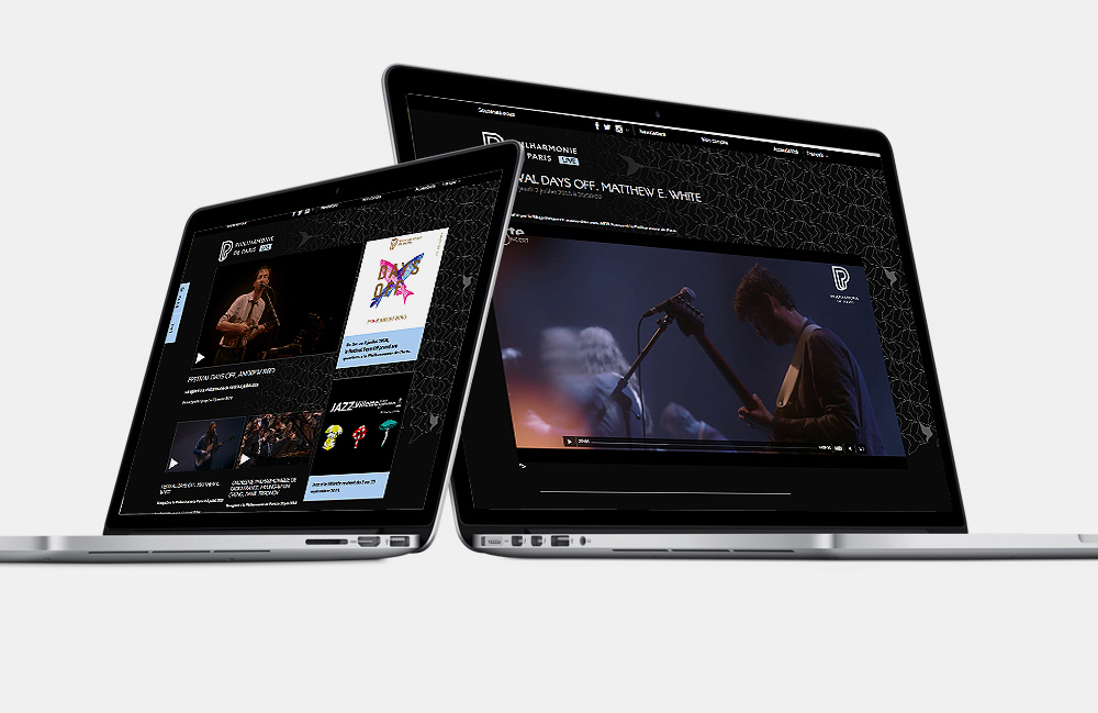 L'image montre deux ordinateurs portables inclinés, affichant des écrans avec ce qui semble être du contenu vidéo et des informations sur un artiste ou un événement musical. L'un des écrans montre un clip ou une performance, tandis que l'autre pourrait contenir des détails supplémentaires ou des panels. Le design des écrans est élégant et moderne, avec un fond noir et une interface stylisée.