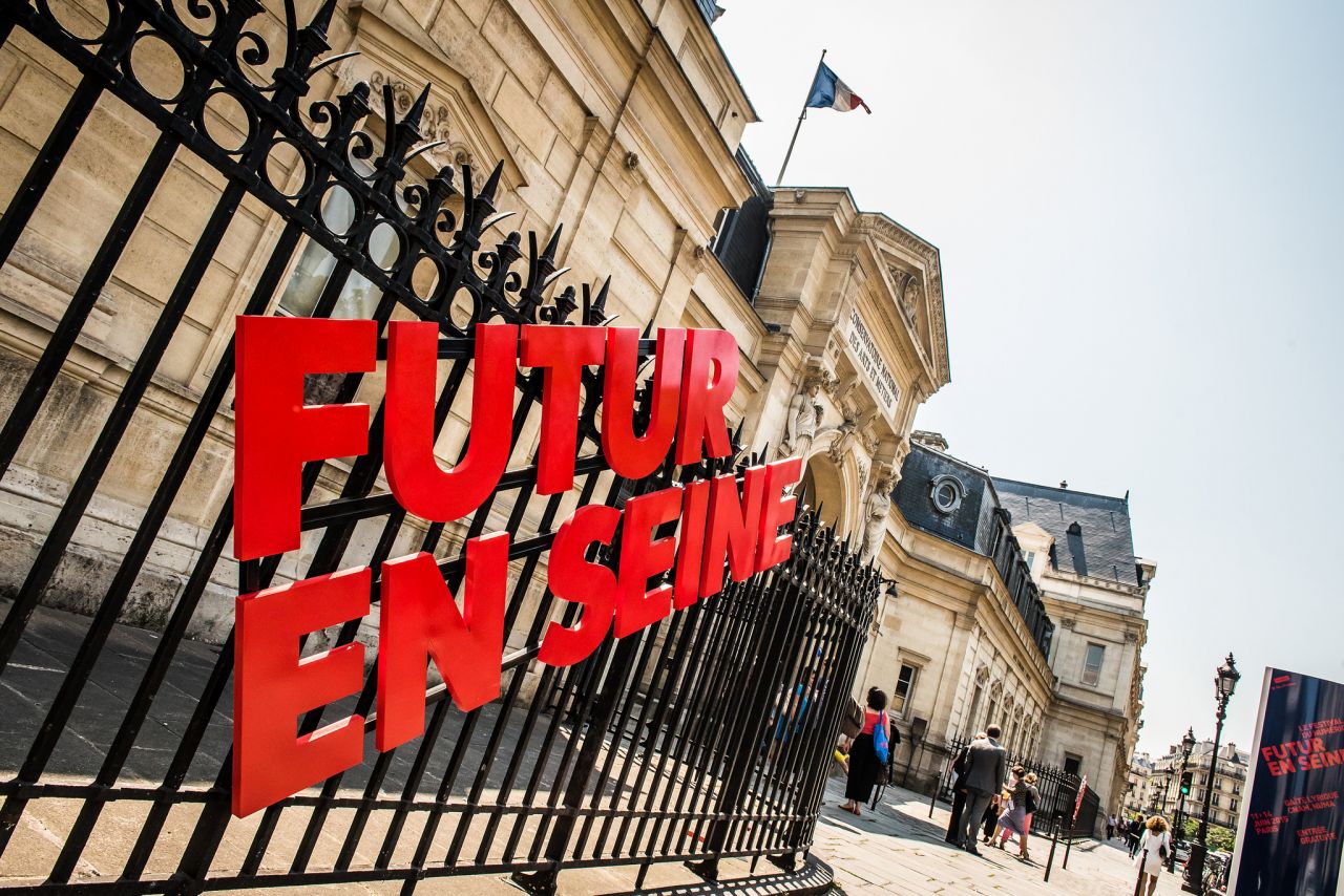 L'image montre un bâtiment, probablement une institution importante, avec un grille en fer forgé. Sur la grille, il y a une inscription en lettres rouges qui dit "FUTUR EN SEINE". En arrière-plan, on aperçoit des gens qui déambulent, et un drapeau est visible sur le toit du bâtiment. Le ciel est clair et ensoleillé, ce qui donne une ambiance sympathique à la scène.