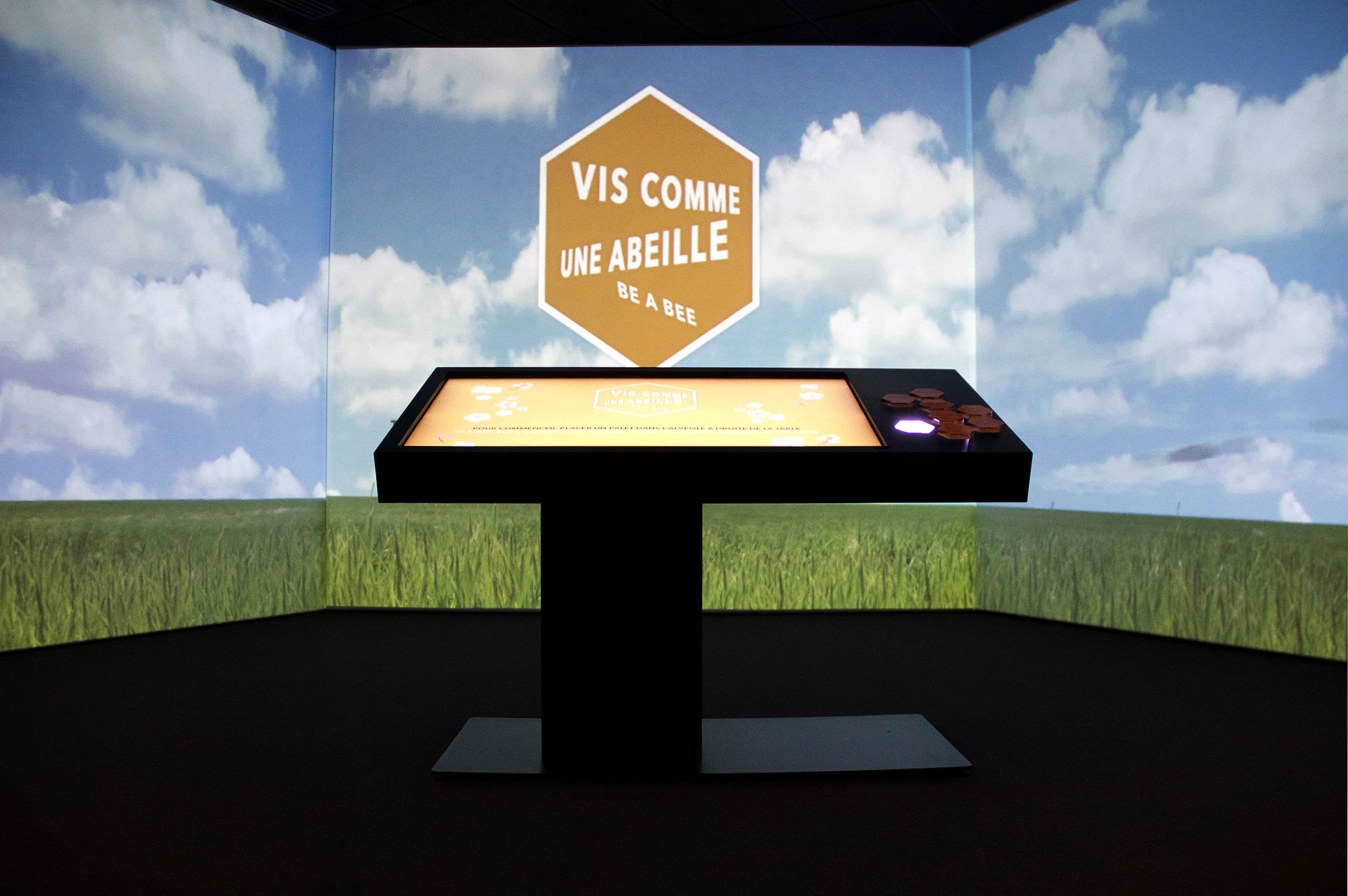 L'image montre une installation interactive dans une salle, avec un écran de projection qui affiche un ciel bleu avec des nuages. Au centre, il y a une table tactile noire sur laquelle se trouve le texte "VIS COMME UNE ABEILLE" en orange, accompagné de la phrase "be a bee". L'environnement dégage une atmosphère ludique et éducative, suggérant un thème lié aux abeilles et à leur comportement. Les murs projetent des images de nature, comme des champs de verdure.