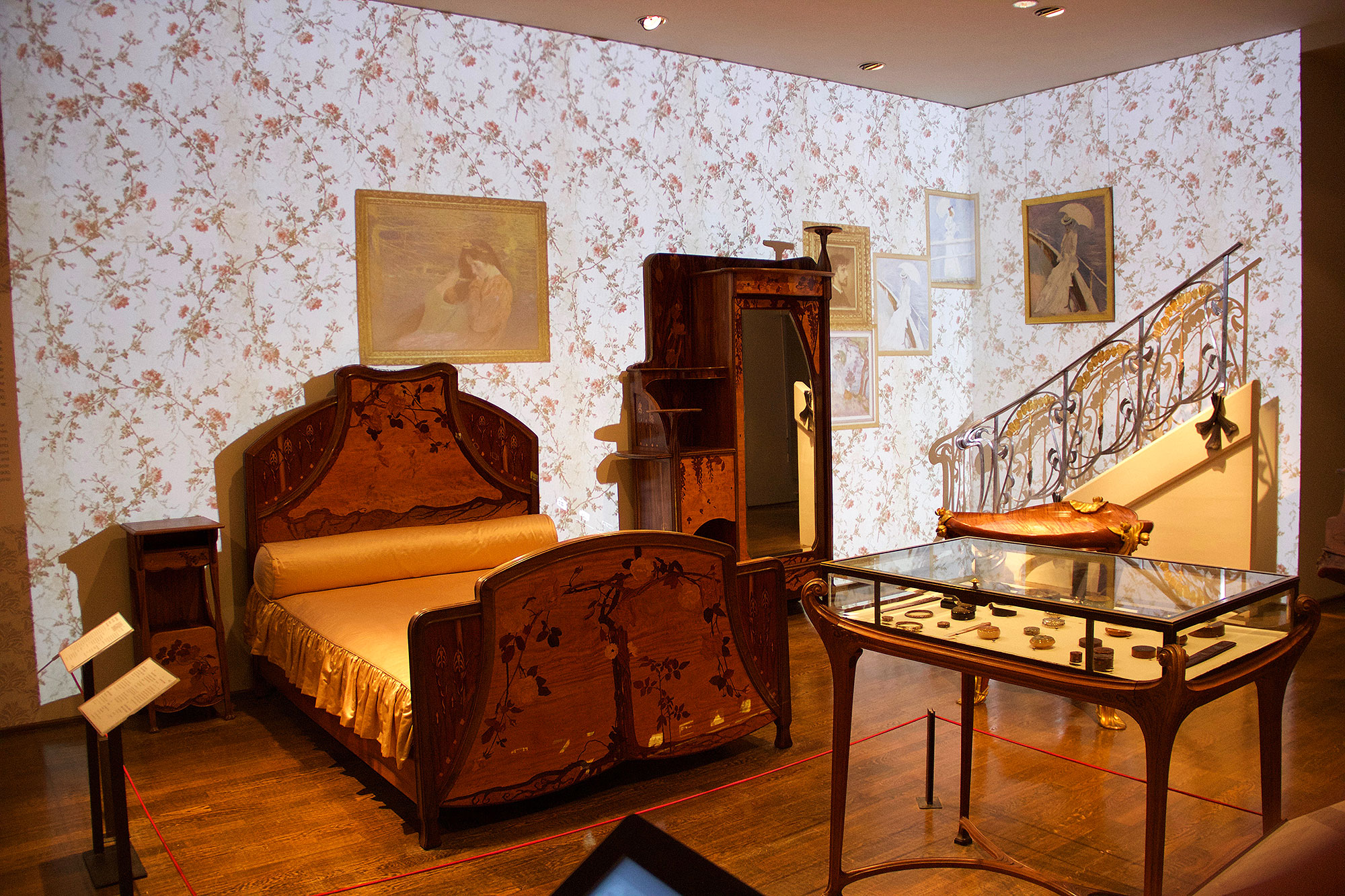 L'image montre une chambre d'époque avec un style décoratif élégant. On y voit un lit en bois orné de motifs floraux, une armoire au design similaire, et une table en verre reposant sur un sol en bois. Les murs sont recouverts d'un papier peint à fleurs, et des tableaux sont accrochés, contribuant à l'ambiance artistique de la pièce. Un escalier avec une balustrade ornée se trouve à l'arrière-plan, ajoutant une touche architecturale à l'ensemble. La lumière douce et les éléments décoratifs évoquent une atmosphère nostalgique et raffinée.