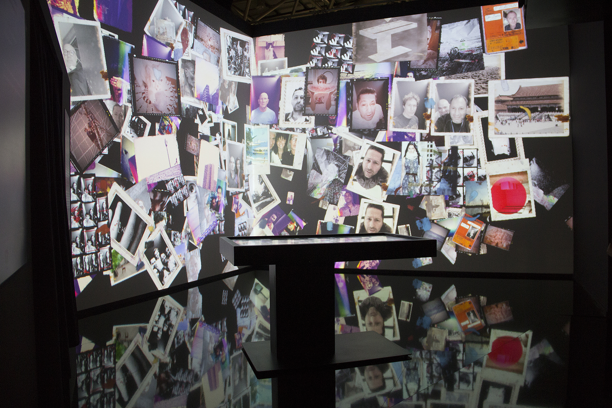 L'image montre une installation artistique immersive où des photographies sont projetées sur les murs. Ces images sont disposées de manière disparate, créant une mosaïque visuelle. On peut voir une variété de photos, dont certaines semblent être des portraits et d'autres des paysages ou des objets. Le sol est noir et réfléchissant, ajoutant une dimension supplémentaire à l'ensemble. L'atmosphère un peu sombre et l'accumulation de ces souvenirs visuels semblent inviter à une réflexion personnelle ou à une exploration du passé.