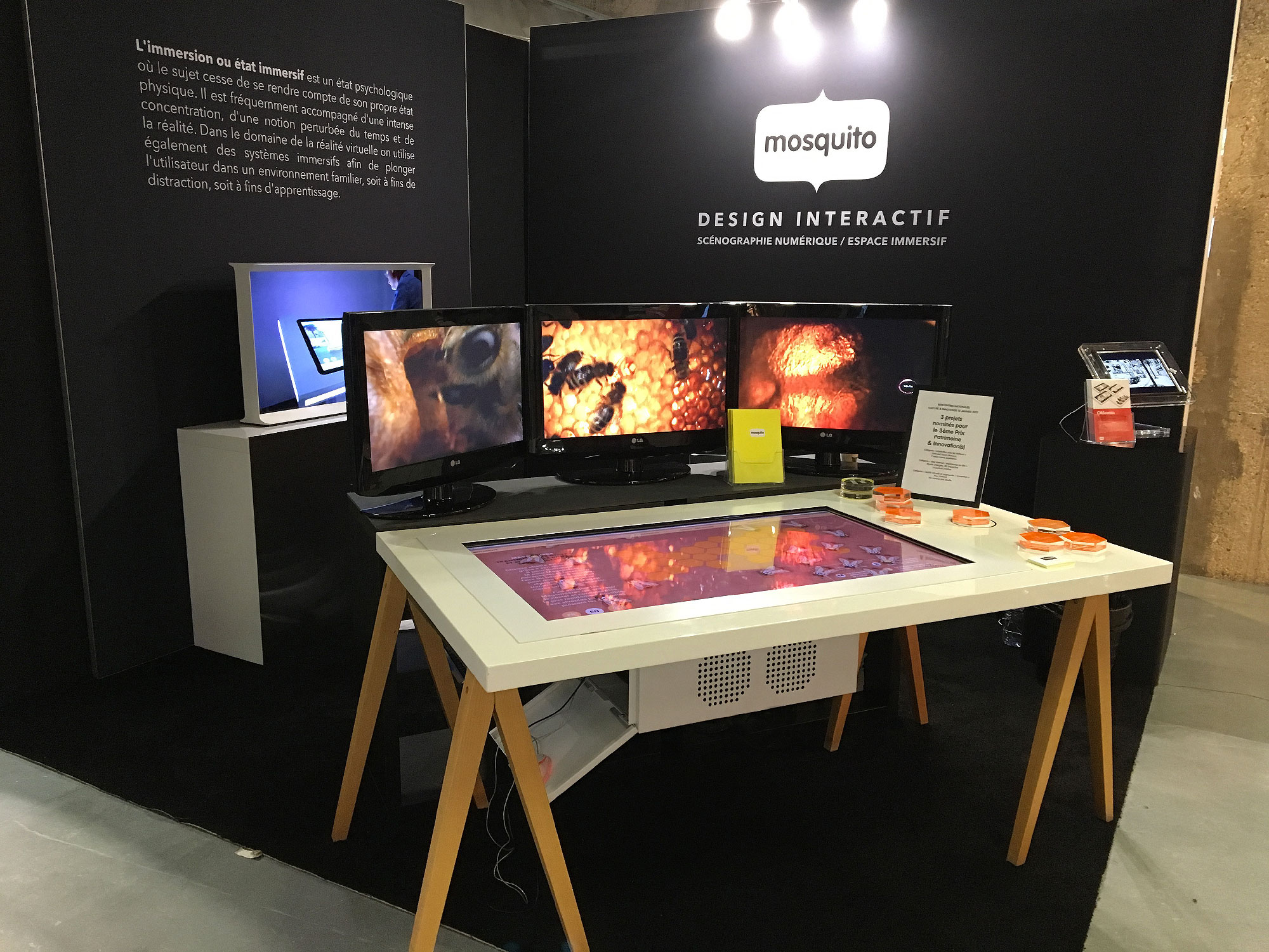 L'image montre un stand interactif nommé "Mosquito", qui semble se concentrer sur une expérience immersive. Au fond, il y a plusieurs écrans affichant des contenus visuels captivants, probablement liés à des thèmes de la nature ou de la technologie. Au centre, une table interactive avec un écran tactile permet aux utilisateurs d'interagir avec le contenu. Des petits objets sont disposés sur la table, qui pourrait encourager une exploration tactile. Le tout est installé dans un environnement sombre, ce qui accentue l'aspect immersif et captivant de la présentation.