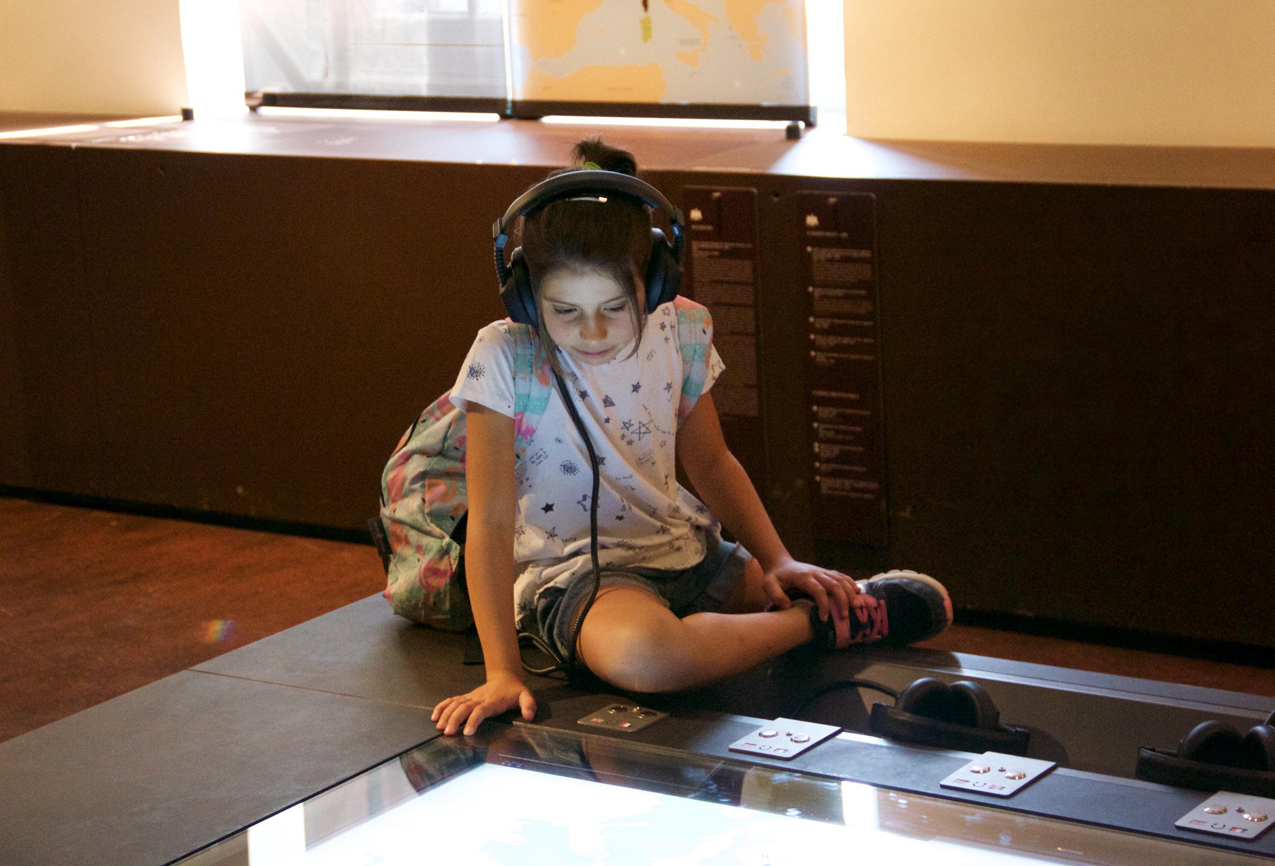 L'image montre une jeune fille assise sur le sol, avec des écouteurs sur les oreilles. Elle porte un t-shirt blanc avec des motifs et un sac à dos coloré. Elle semble concentrée sur un écran interactif devant elle, qui pourrait afficher une carte ou une information. L'environnement est celui d'un musée, avec un éclairage naturel et des panneaux explicatifs en arrière-plan.