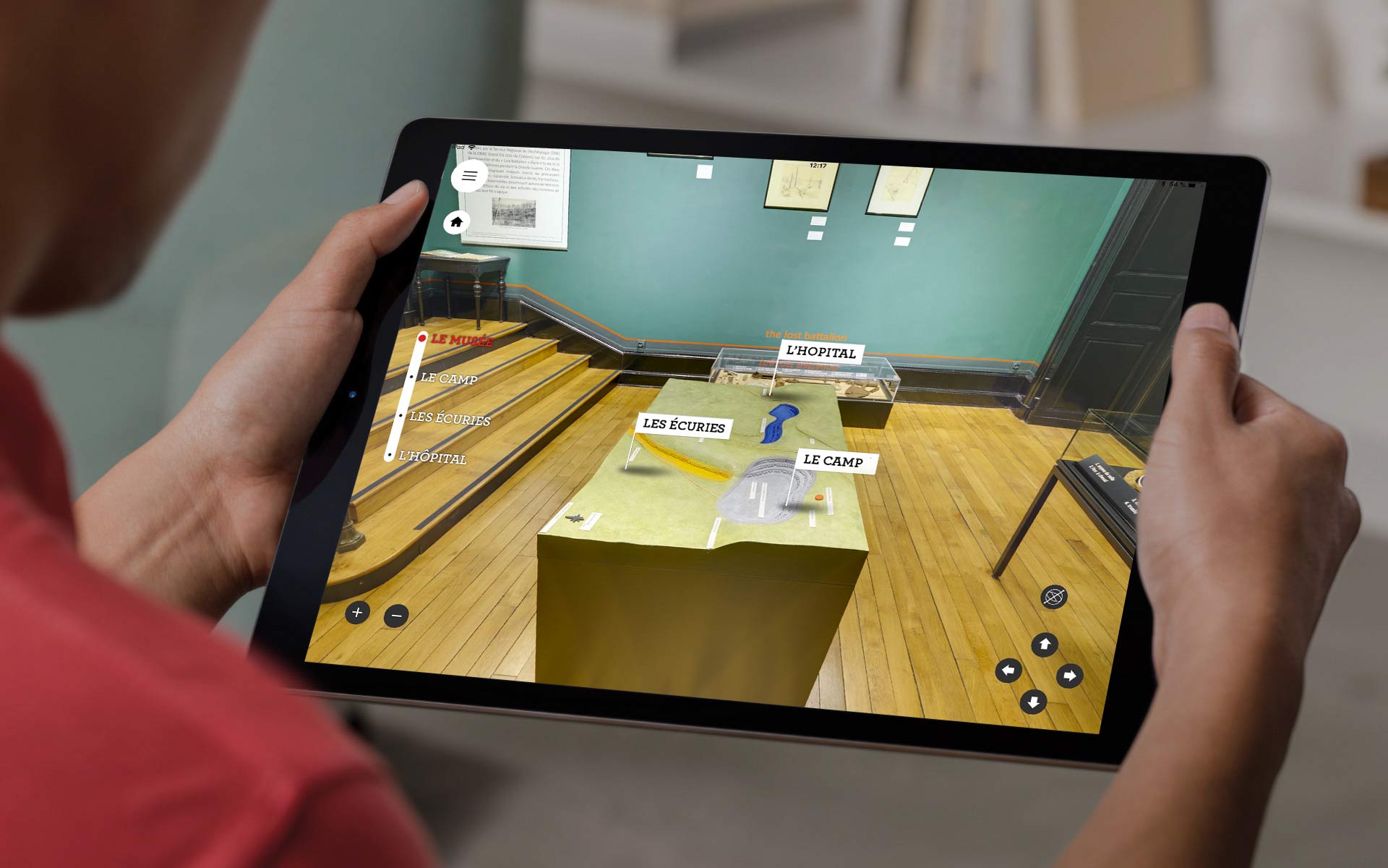 L'image montre une personne tenant une tablette, sur laquelle est affichée une application de réalité augmentée. Sur l'écran, on peut voir des éléments interactifs en rapport avec une exposition, avec des étiquettes comme "Les découvertes" et "Le camp". Le fond de l'image représente un musée, avec des murs verts et un sol en bois. La personne semble explorer des informations ou des objets exposés, mettant en avant l'utilisation de la technologie pour enrichir l'expérience muséale.