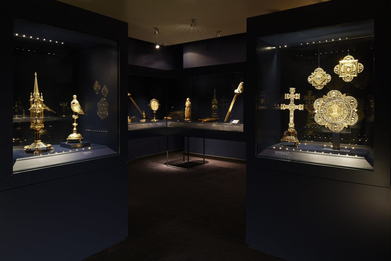 L'image montre une salle d'exposition dans un musée, mettant en valeur plusieurs objets en or. On peut voir des vitrines bien éclairées contenant des pièces de décoration, probablement des artefacts religieux ou historiques, comme une croix, des sculptures, et d'autres ornements raffinés. L'éclairage crée une ambiance sophistiquée, mettant en avant la beauté et le détail des pièces exposées. Les murs sont sombres, ce qui accentue le brillant des objets en or.