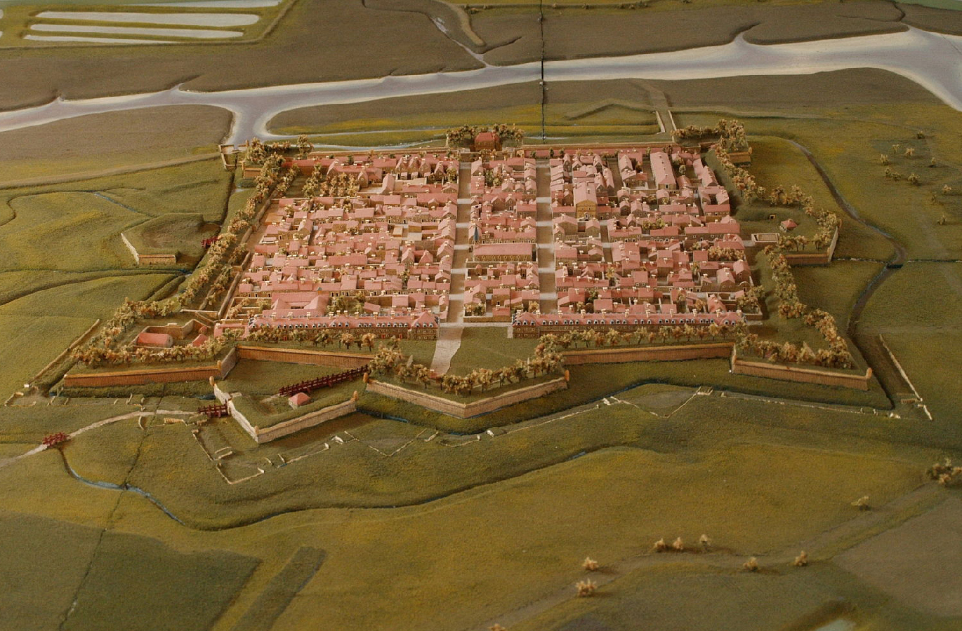 L'image montre un modèle réduit d'une ville fortifiée, probablement d'une époque historique. On y voit une disposition ordonnée avec des rues, des bâtiments et des espaces verts. La ville est entourée de remparts, suggérant qu'elle était défendue contre d'éventuelles attaques. Des canaux ou des rivières sont visibles à proximité, indiquant une utilisation stratégique de l'eau. L'ensemble dénote une planification minutieuse et un souci du détail dans l'architecture de l'époque.