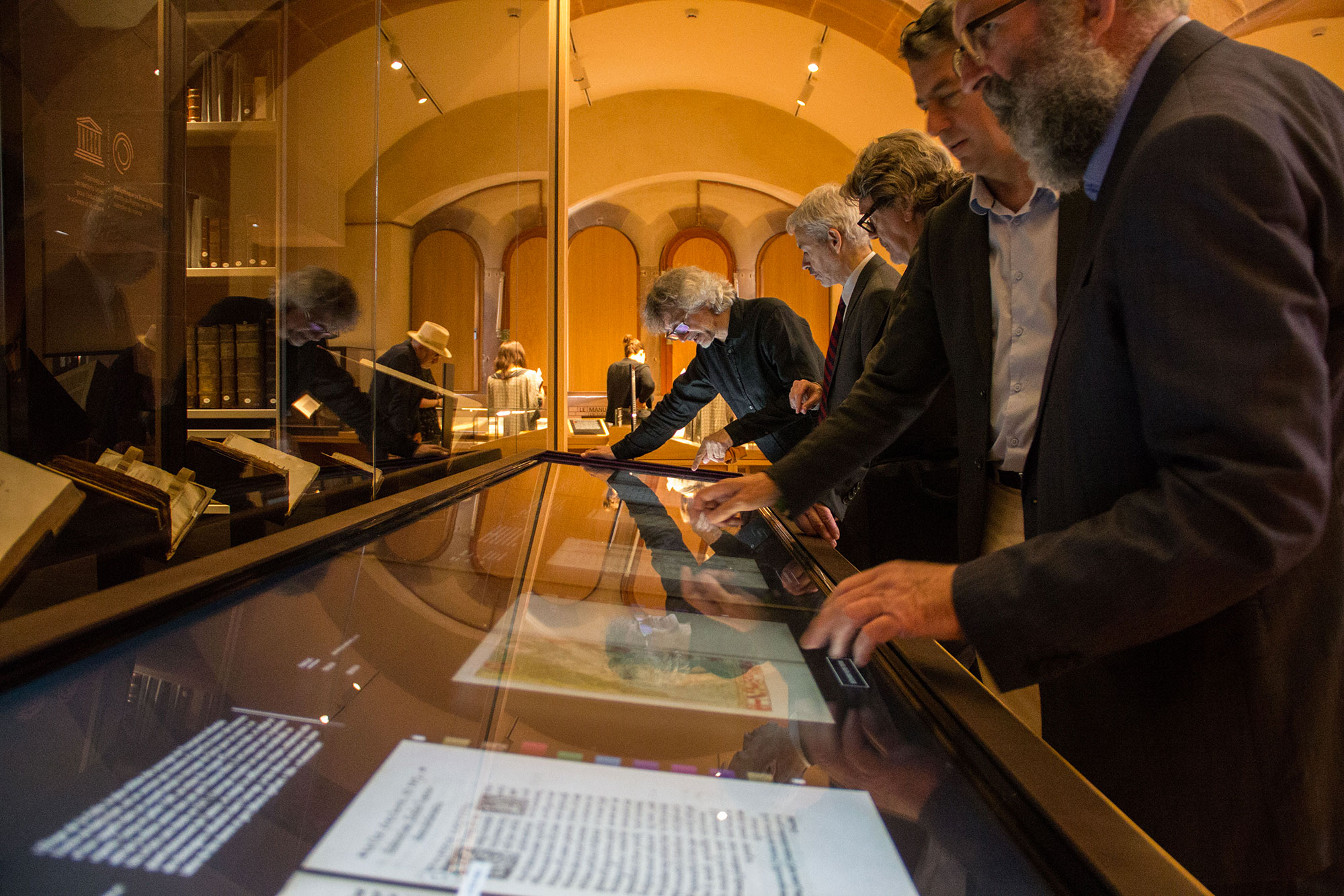 L'image montre un groupe de personnes debout devant une exposition dans un musée. Elles semblent intéressées et observent des objets présentés sur une table interactive. Les visiteurs, de différents âges et styles vestimentaires, touchent l'écran pour explorer les informations ou les images. En arrière-plan, on aperçoit des livres et des documents affichés dans des vitrines. L'éclairage est doux, créant une ambiance chaleureuse et propice à la découverte.