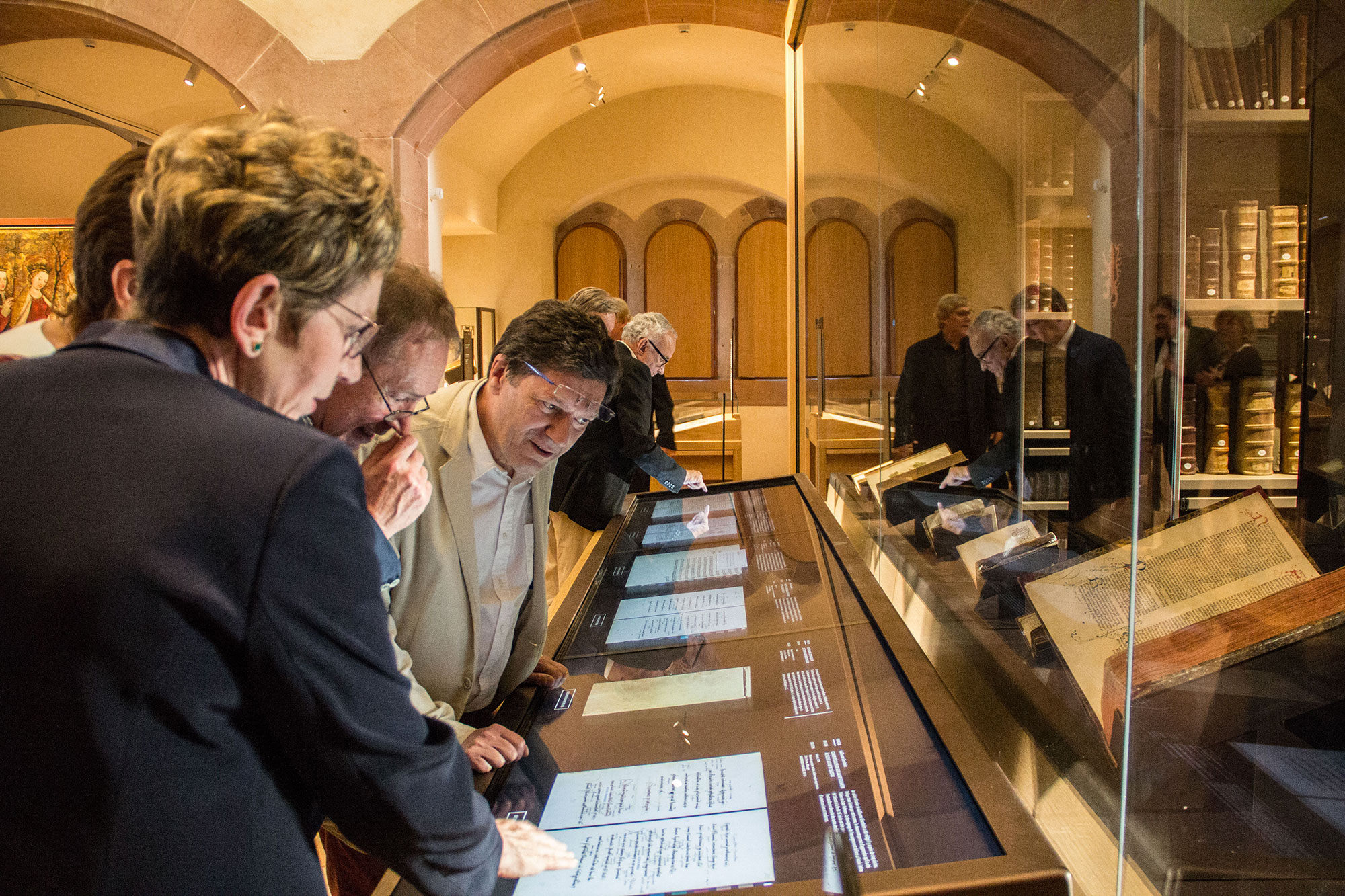 L'image montre un groupe de personnes réunies autour d'une vitrine dans un musée. Ils examinent des documents ou des manuscrits exposés, probablement de grande valeur historique. Les visiteurs semblent intéressés et engagés dans leur découverte, avec des expressions de curiosité. La lumière de l'espace muséal met en avant les objets exposés, créant une atmosphère d'exploration et d'apprentissage. En arrière-plan, d'autres personnes floues sont également présentes, profitant de l'exposition.