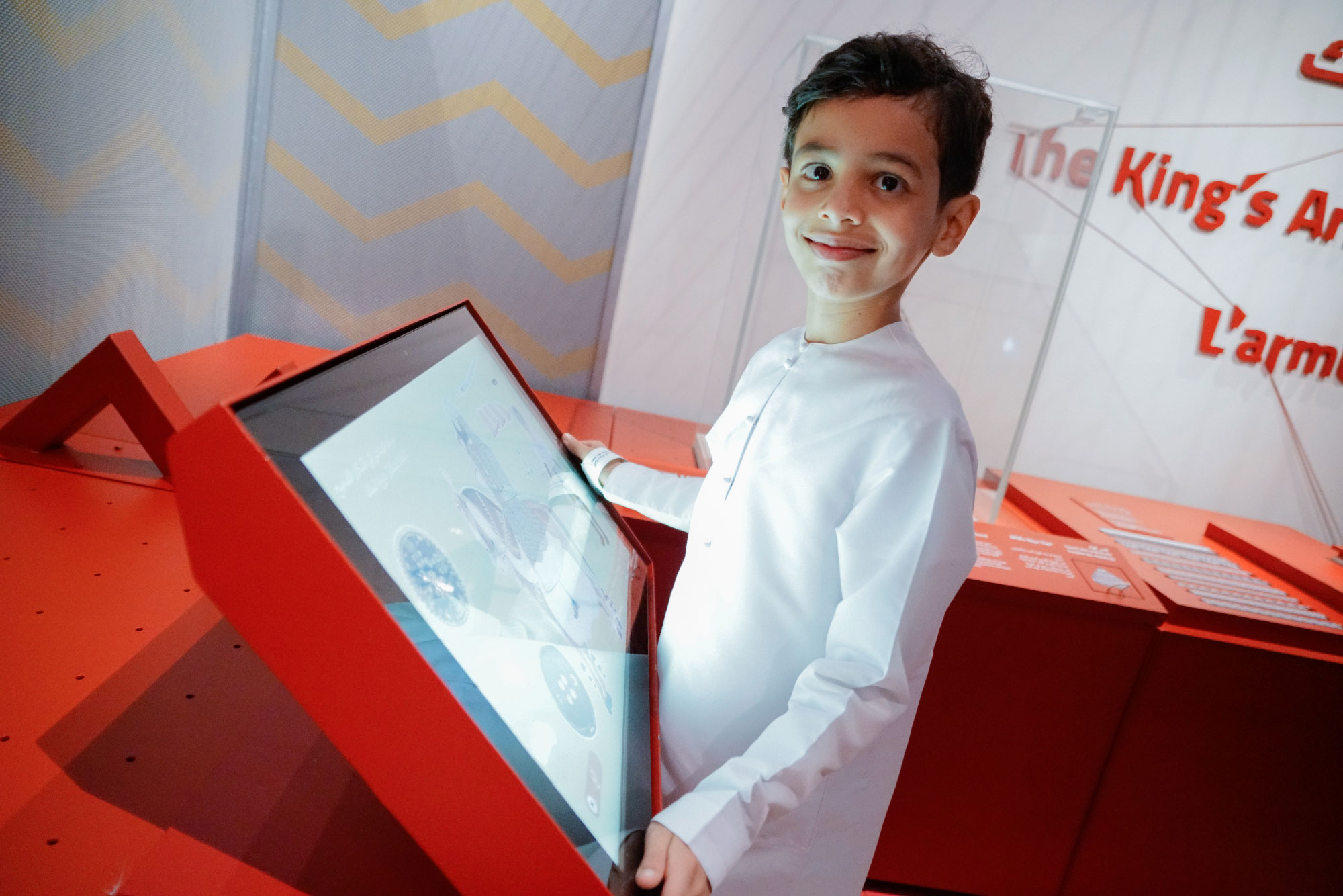 L'image montre un jeune garçon souriant, habillé d'une tenue traditionnelle blanche. Il se tient devant un écran interactif dans un environnement exposé, probablement dans un musée. L'interface de l'écran semble afficher des informations ou des graphiques. L'arrière-plan est minimaliste, avec des formes géométriques et des couleurs vives, principalement du rouge.