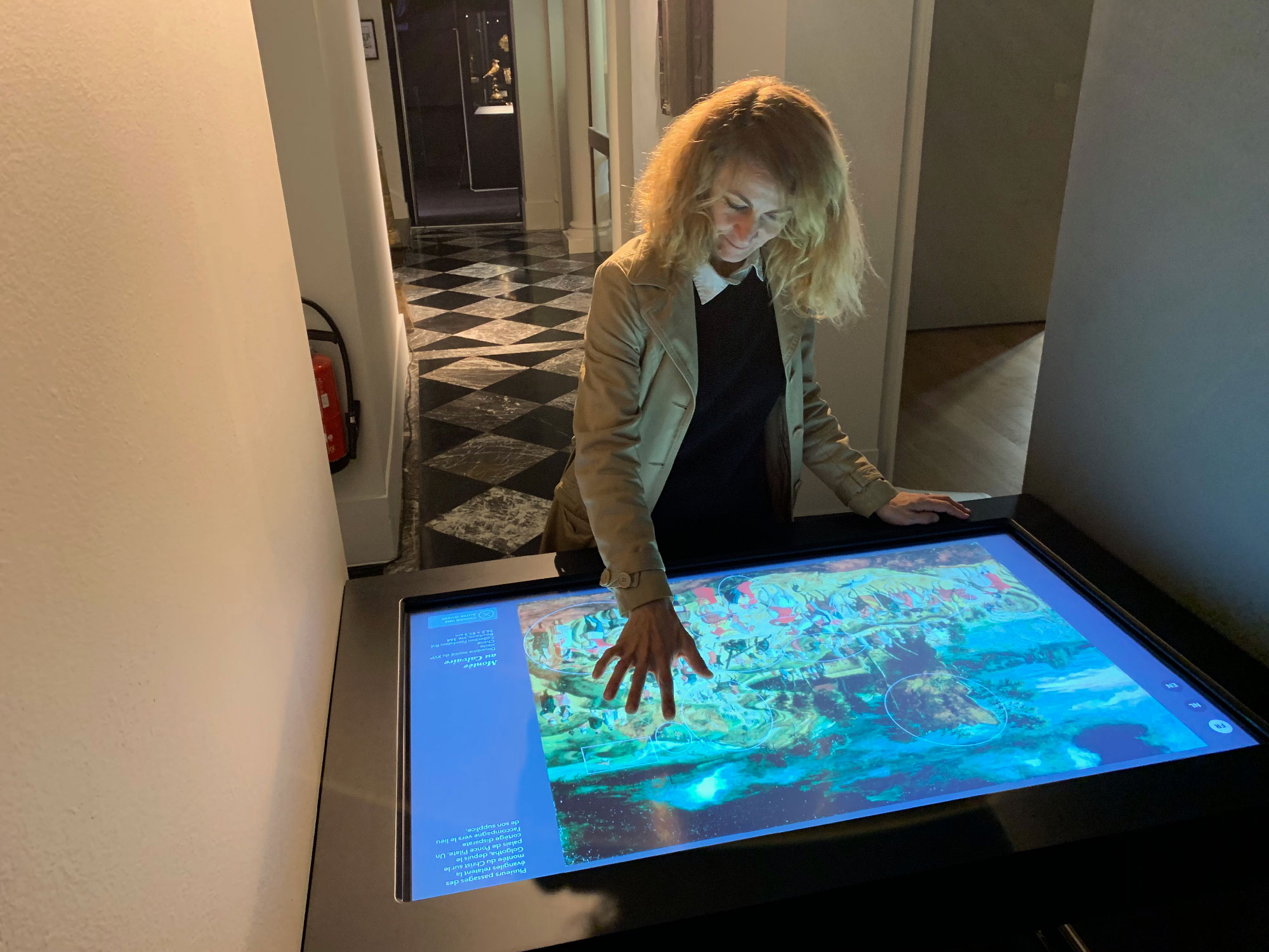 Sur l'image, on voit une femme aux cheveux blonds bouclés qui interagit avec un écran tactile. Elle semble examinée une œuvre d'art ou une illustration colorée affichée sur la surface de la table. L'environnement est élégant, avec un sol en damier noir et blanc, et on aperçoit des murs clairs. Dans le fond, des portes et des couloirs suggèrent un espace muséal. La scène dégage une atmosphère de curiosité et d'exploration.