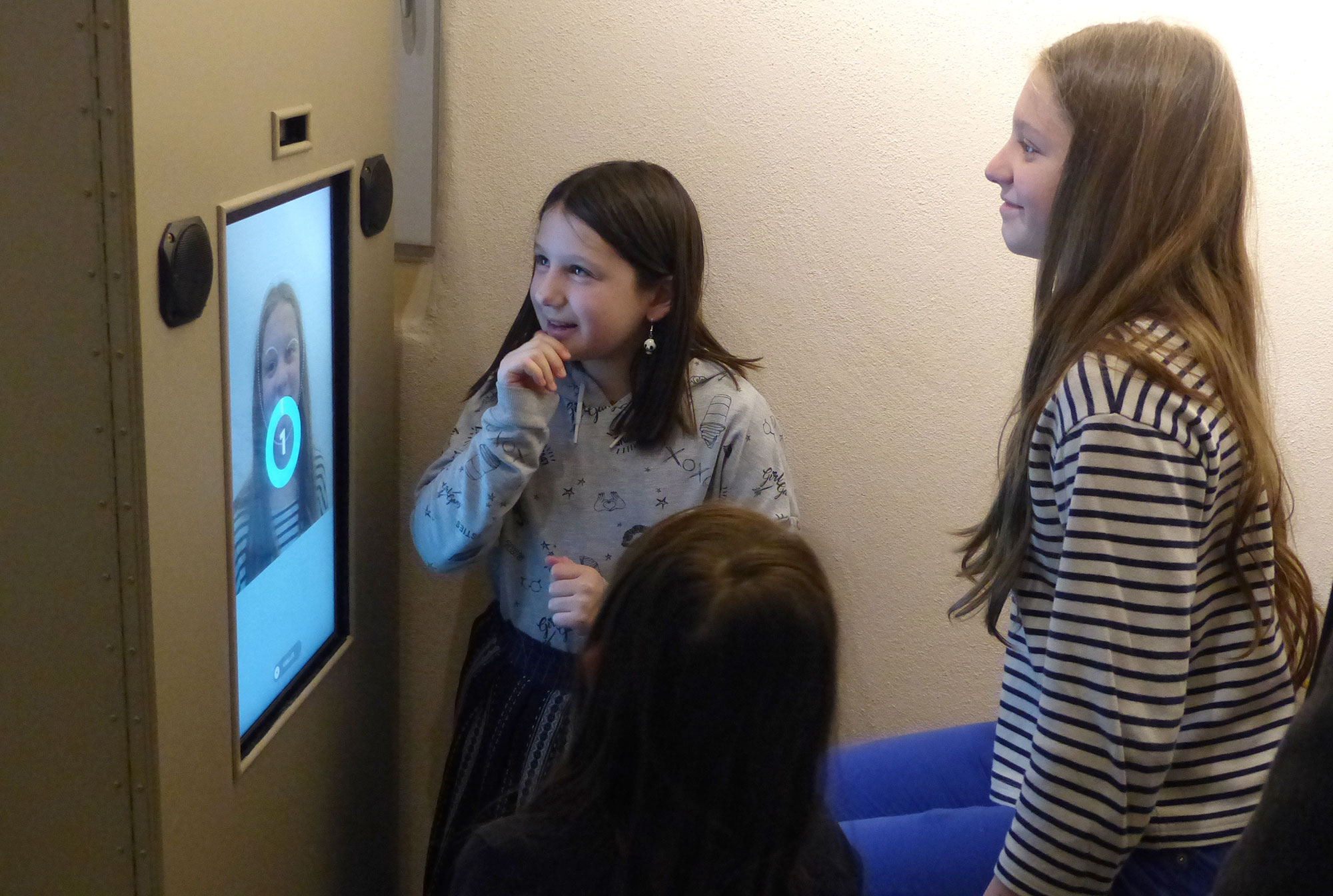 L'image montre un groupe de jeunes filles interagissant avec un écran. L'une d'elles, qui se tient devant l'écran, semble surprise ou amusée, tandis que les autres regardent avec intérêt. L'écran affiche une image qui pourrait être un reflet ou une vidéo d'elles. L'ambiance semble conviviale et ludique. Les filles portent des vêtements décontractés, ce qui renforce l'impression d'un moment informel et joyeux.