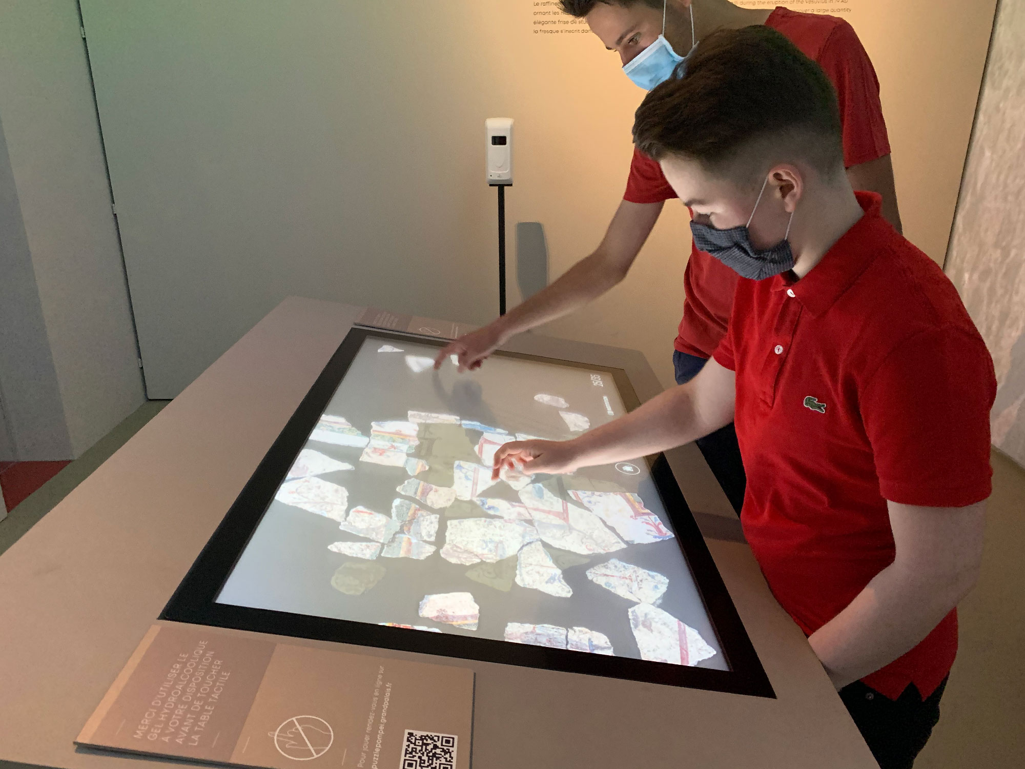 L'image montre deux jeunes garçons en train d'interagir avec un panneau tactile. Ils portent des masques et sont habillés en t-shirts rouges. Le panneau affiche des images de morceaux de pierres ou de fragments, probablement dans un contexte éducatif ou muséal, où ils explorent et découvrent des informations. L'ambiance semble moderne et engageante, favorisant l'apprentissage interactif.