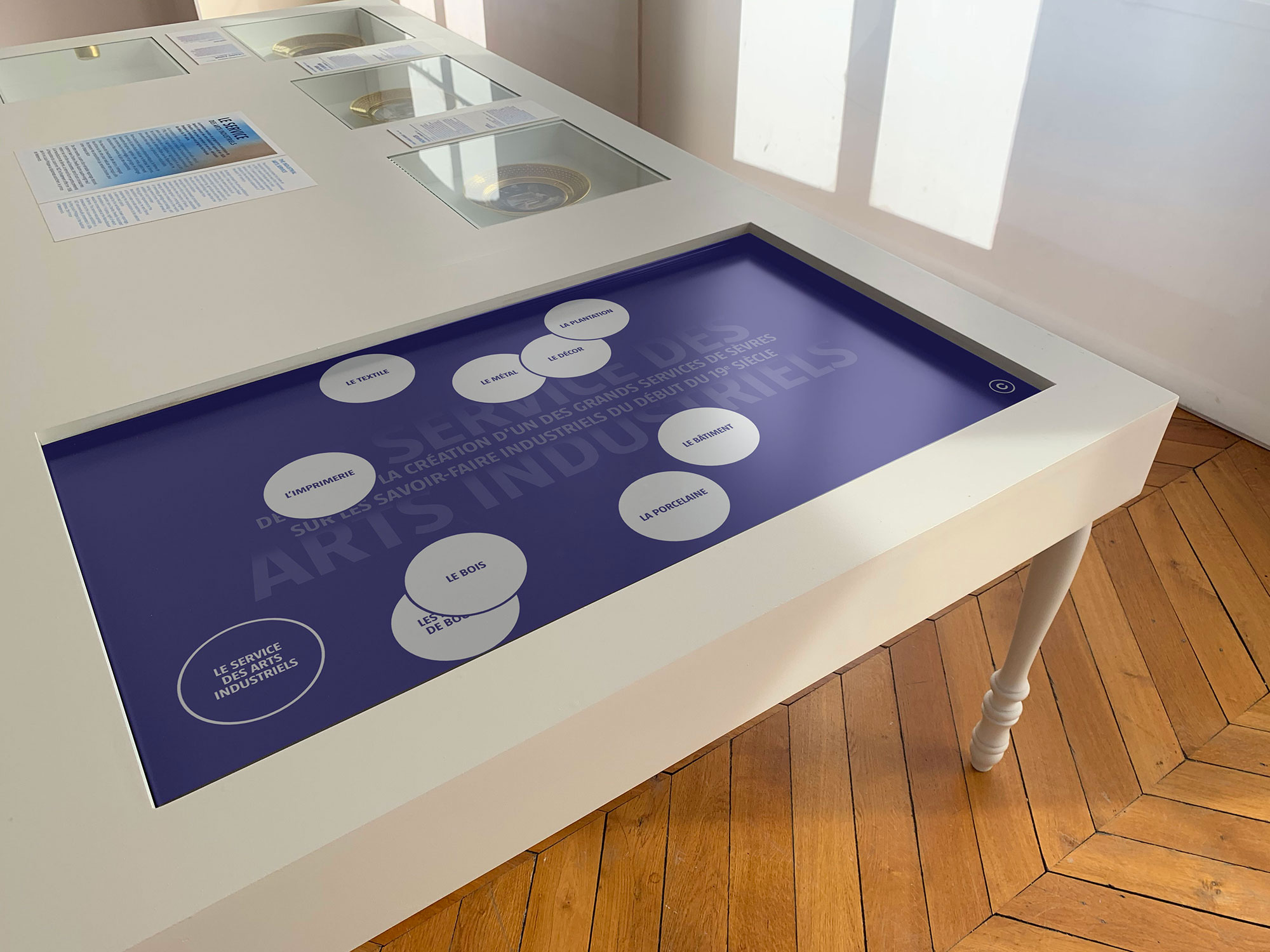 L'image montre une table d'exposition design moderne avec une surface interactive. La table semble être utilisée dans un contexte muséal ou éducatif. Elle possède des éléments ronds et des illustrations ou textes disposés autour d'un écran tactile au centre. Le fond de l'écran est de couleur violette et inclut des éléments graphiques liés aux arts industriels. La table a des pieds en bois, et le sol est en parquet. Les murs environnants semblent clairs, ce qui donne une atmosphère lumineuse à l'espace.
