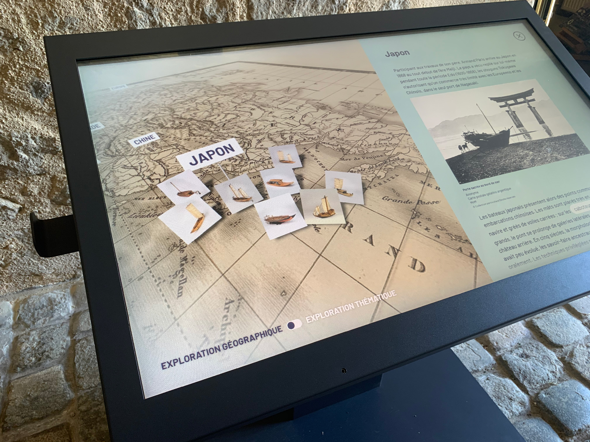 L'image montre un écran tactile interactif présentant une carte géographique du Japon, avec des éléments disposés sur cette carte. On peut voir des informations textuelles sur le Japon sur le côté droit de l'écran, ainsi que des icônes représentant des objets ou thèmes de l'exploration. Le fond de l'écran a un motif qui ressemble à une carte ancienne. Le tout semble être installé dans un environnement muséal avec un sol en pierre.