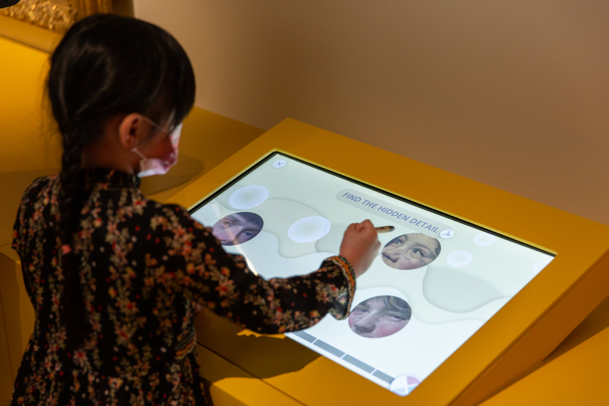 L'image montre une jeune fille utilisant un écran tactile. Elle semble concentrée sur une activité interactive, où elle doit trouver un détail caché. L'écran affiche des visages flous ou modifiés, et la représentation de l'interface utilise des formes fluides. La fille porte un masque et est habillée d'une robe à motifs floraux, ce qui ajoute une touche colorée à la scène. Le décor est minimaliste, avec un fond jaune qui attire l'attention sur l'écran.