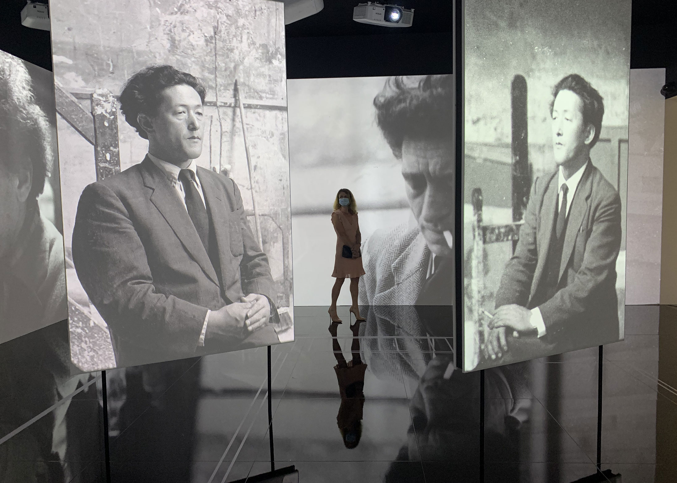 L'image présente une installation artistique moderne où une silhouette féminine se tient au centre d'un espace d'exposition. Cette personne porte un masque et se trouve entre plusieurs écrans qui affichent des photographies en noir et blanc. Ces images montrent un homme dans des poses réfléchies, évoquant une ambiance nostalgique et contemplative. La lumière des projections crée des reflets sur le sol, ajoutant une dimension dynamique à l'ensemble de la scène.