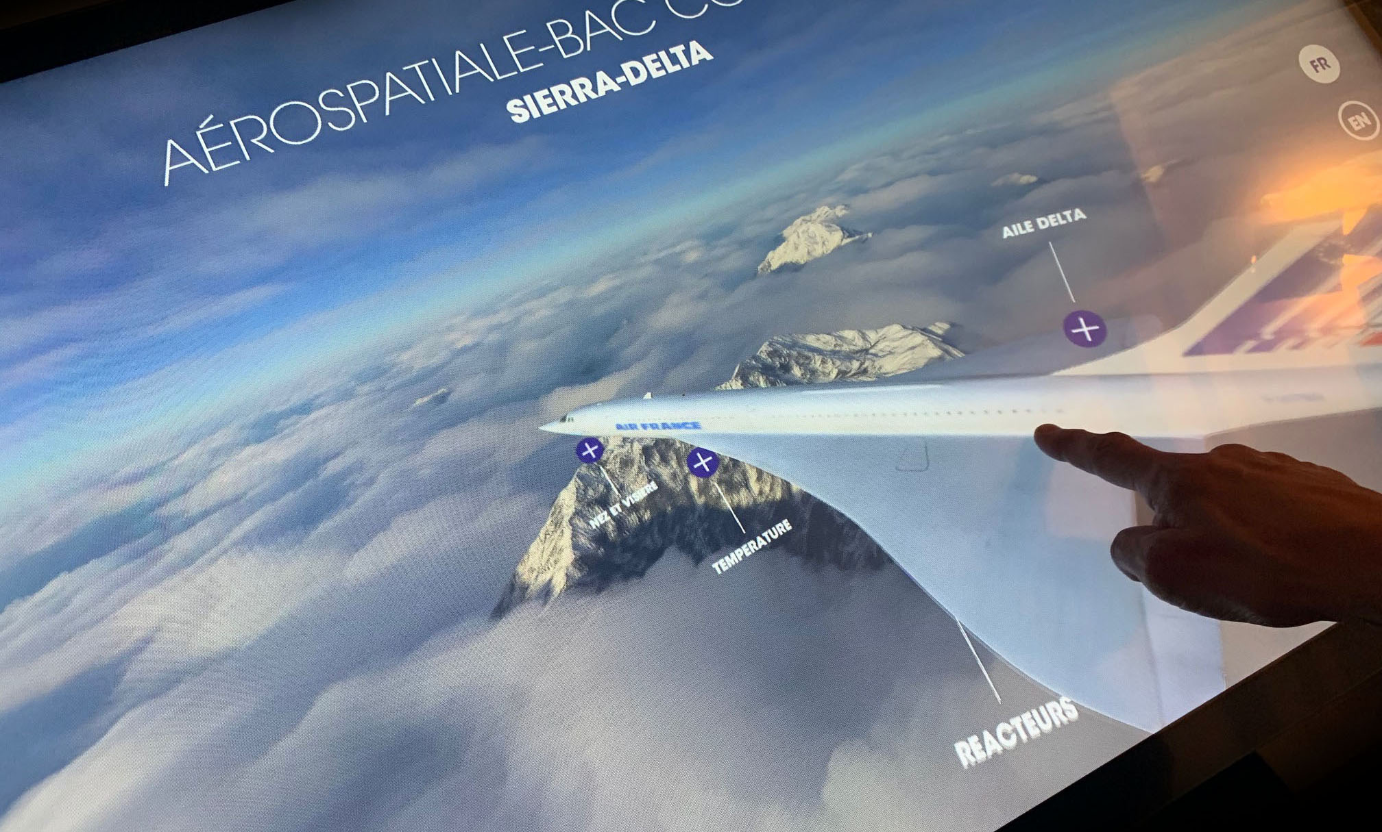 L'image montre un écran interactif qui présente un modèle d'avion dans un environnement aérien. On peut voir des montagnes sous des nuages, et un doigt pointe vers l'aile de l'appareil, où des annotations étiquettent des éléments comme "Aile Delta", "Réacteur" et "Température". Le titre indique qu'il s'agit d'une présentation sur l'aéronautique, plus précisément sur le modèle "Sierra-Delta".