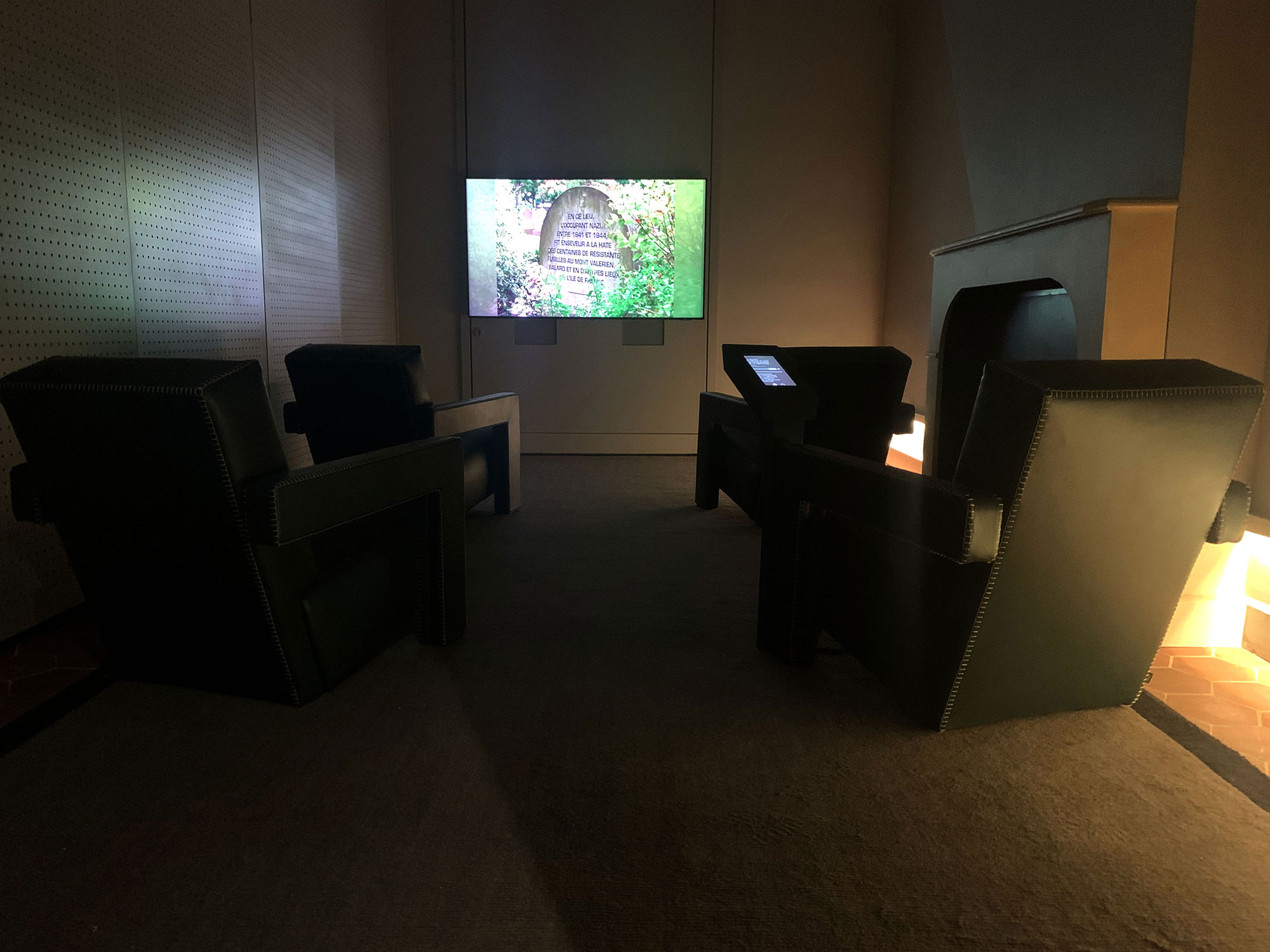 L'image montre une pièce avec un éclairage tamisé. Au centre, il y a quatre fauteuils en cuir noir disposés face à un écran de télévision, où une image de marches entourées de verdure est affichée. La décoration semble moderne et minimaliste, et le sol est couvert d'un tapis sombre. Les murs sont clairs, tandis qu'une cheminée est visible sur le côté, ajoutant une touche chaleureuse à l'espace.