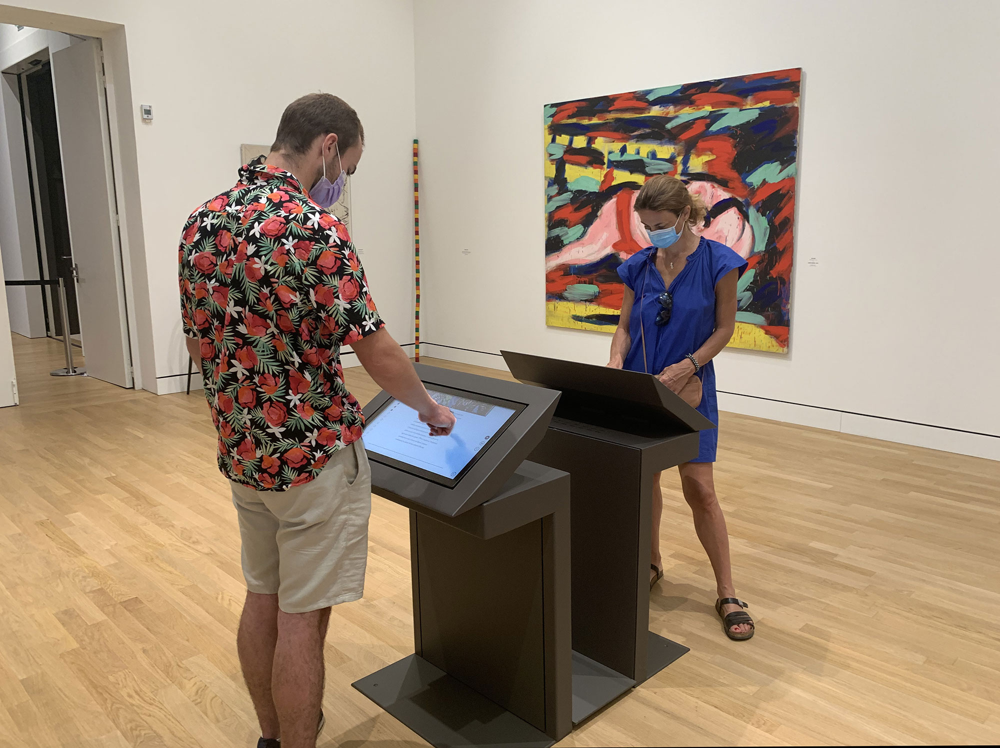 L'image montre deux personnes dans un musée ou une galerie d'art. L'une d'elles, un homme portant une chemise à motifs floraux, est en train d'interagir avec un écran tactile. L'autre personne, une femme en robe bleue, observe ce qu'il fait. Les deux portent des masques de protection. En arrière-plan, on peut voir une toile colorée accrochée au mur, ajoutant une ambiance artistique à l'espace. Le sol est en bois clair.