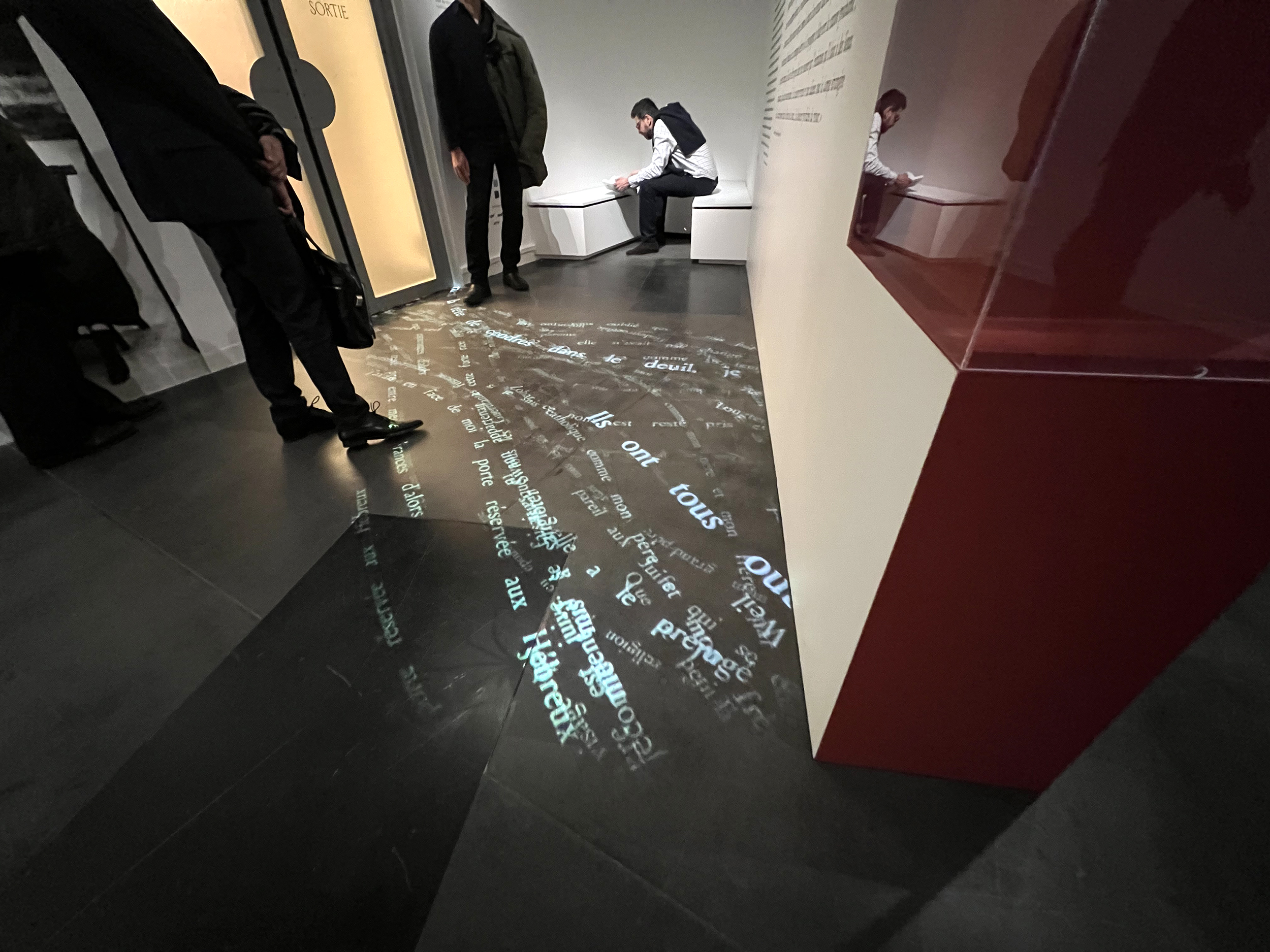 L'image montre un espace intérieur, probablement un musée ou une galerie. Au sol, des mots et des phrases sont projetés, créant un motif lumineux sur le sol. On peut voir des silhouettes de personnes, certaines debout, d'autres assises, interagissant avec l'environnement. Les murs semblent comporter des inscriptions ou des informations, mais elles ne sont pas clairement visibles dans l'image. L'atmosphère semble être moderne et intellectuelle, réfléchissant une intention artistique ou informative.