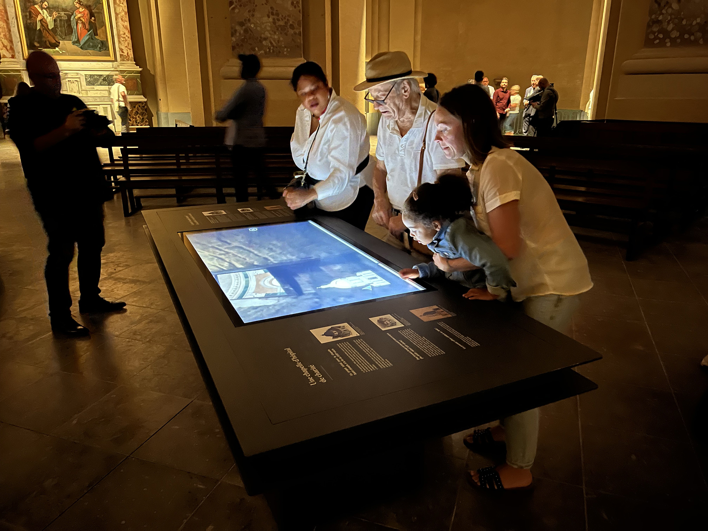 L'image montre un groupe de quatre personnes en train d'observer une table interactive au musée ou dans un espace d'exposition. On peut voir deux adultes et un enfant, concentrés sur l'écran qui affiche probablement des informations ou des images. Une ambiance de curiosité et d'engagement se dégage, alors que d'autres visiteurs se déplacent en arrière-plan. L'éclairage et le décor semblent suggérer un lieu culturel ou historique.