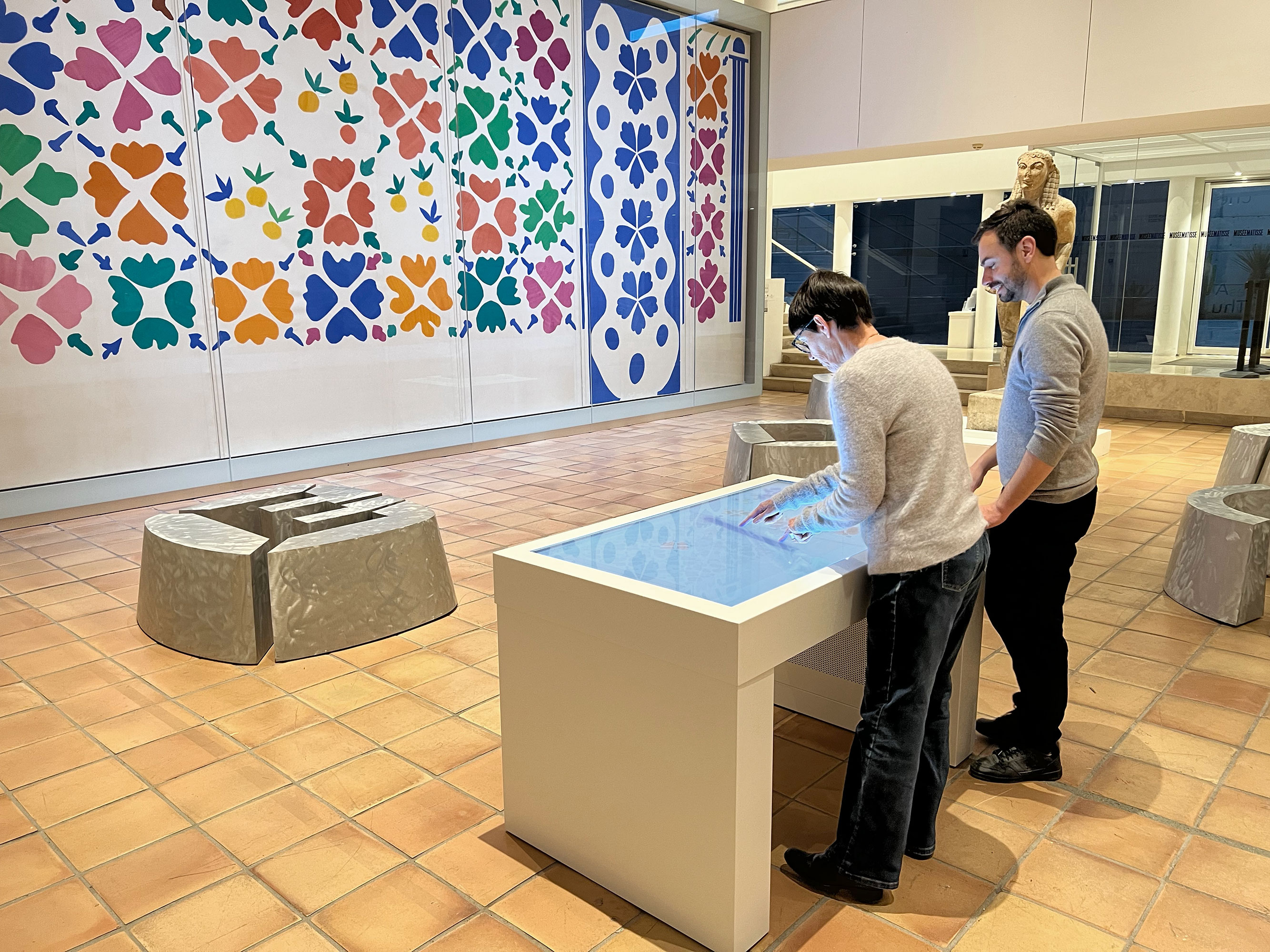 L'image montre une salle d'exposition d'un musée. Deux personnes se tiennent devant un écran interactif en touchant son interface. En arrière-plan, un grand mur est décoré de motifs colorés en forme de fleurs et de feuilles. Il y a également des sculptures ou des objets artistiques disposés dans la salle. Le sol est carrelé avec des carreaux en terre cuite, ajoutant une atmosphère chaleureuse à l'environnement.