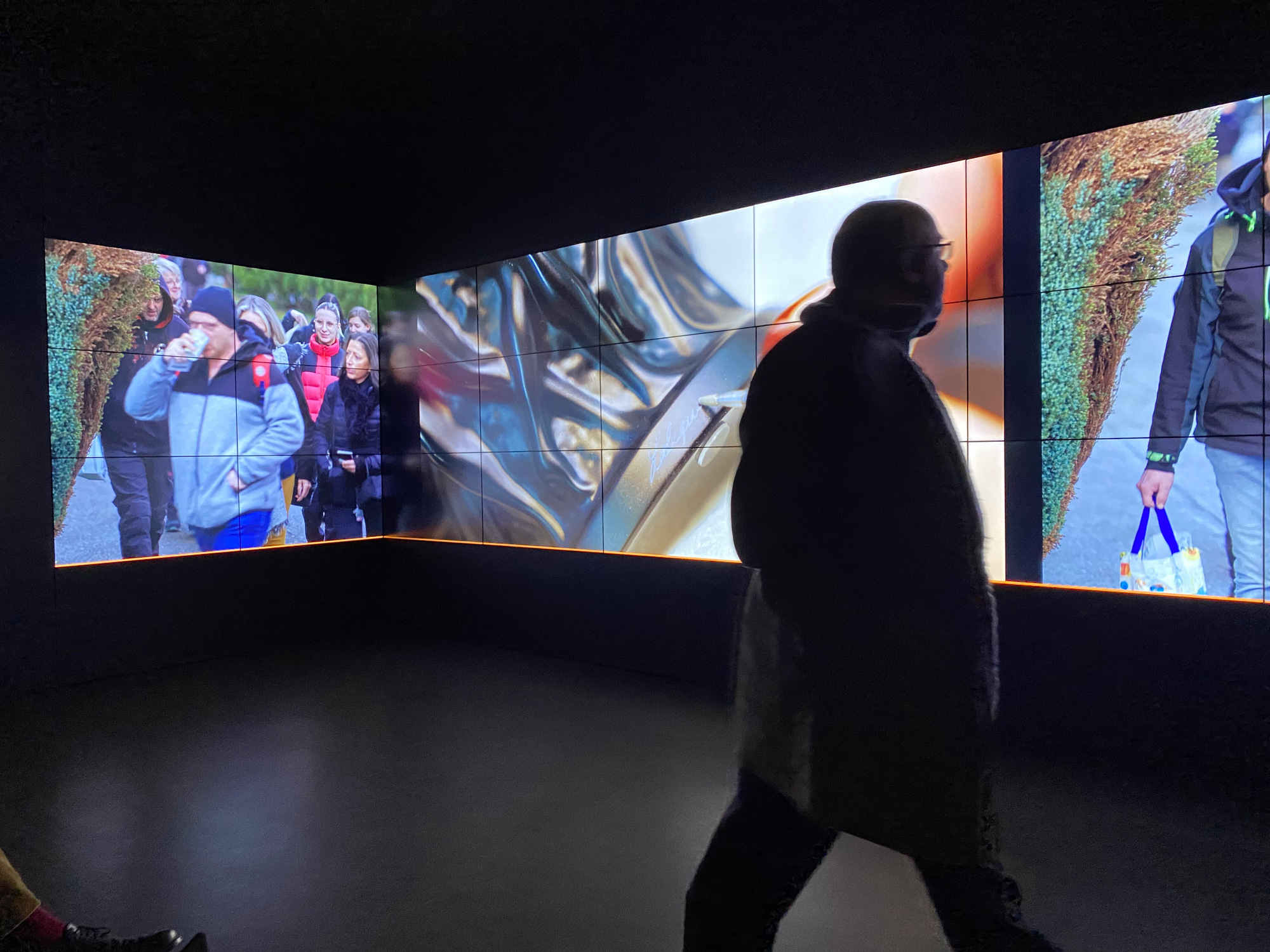 L'image montre un espace intérieur avec plusieurs écrans affichant des images variées. Sur l'un des écrans, on peut apercevoir des personnes en train de marcher, tandis que d'autres écrans présentent des motifs abstraits. Au premier plan, une silhouette d'une personne marche, créant un contraste avec les images dynamiques en arrière-plan. L'éclairage et la disposition des écrans ajoutent une atmosphère moderne et immersive à la scène.