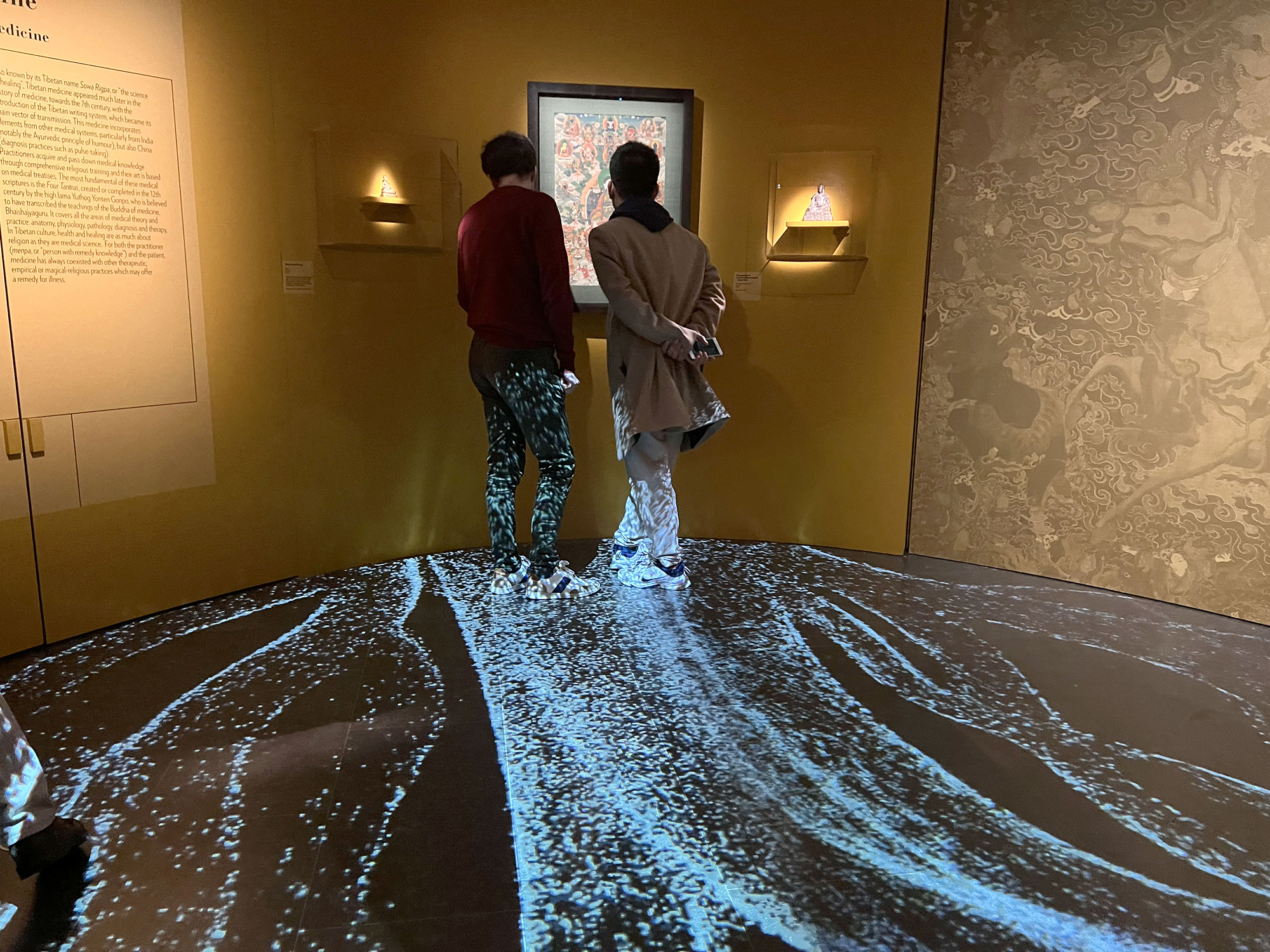 L'image montre deux personnes se tenant debout dans un espace d'exposition. Ils regardent attentivement une œuvre d'art affichée sur le mur. Le sol est recouvert d'une projection qui crée un effet visuel évoquant de l'eau ou des vagues. Les murs sont de couleur jaune, et il y a des éclairages discret sur les côtés qui mettent en valeur l'exposition. L'ambiance est à la fois contemplative et immersive.