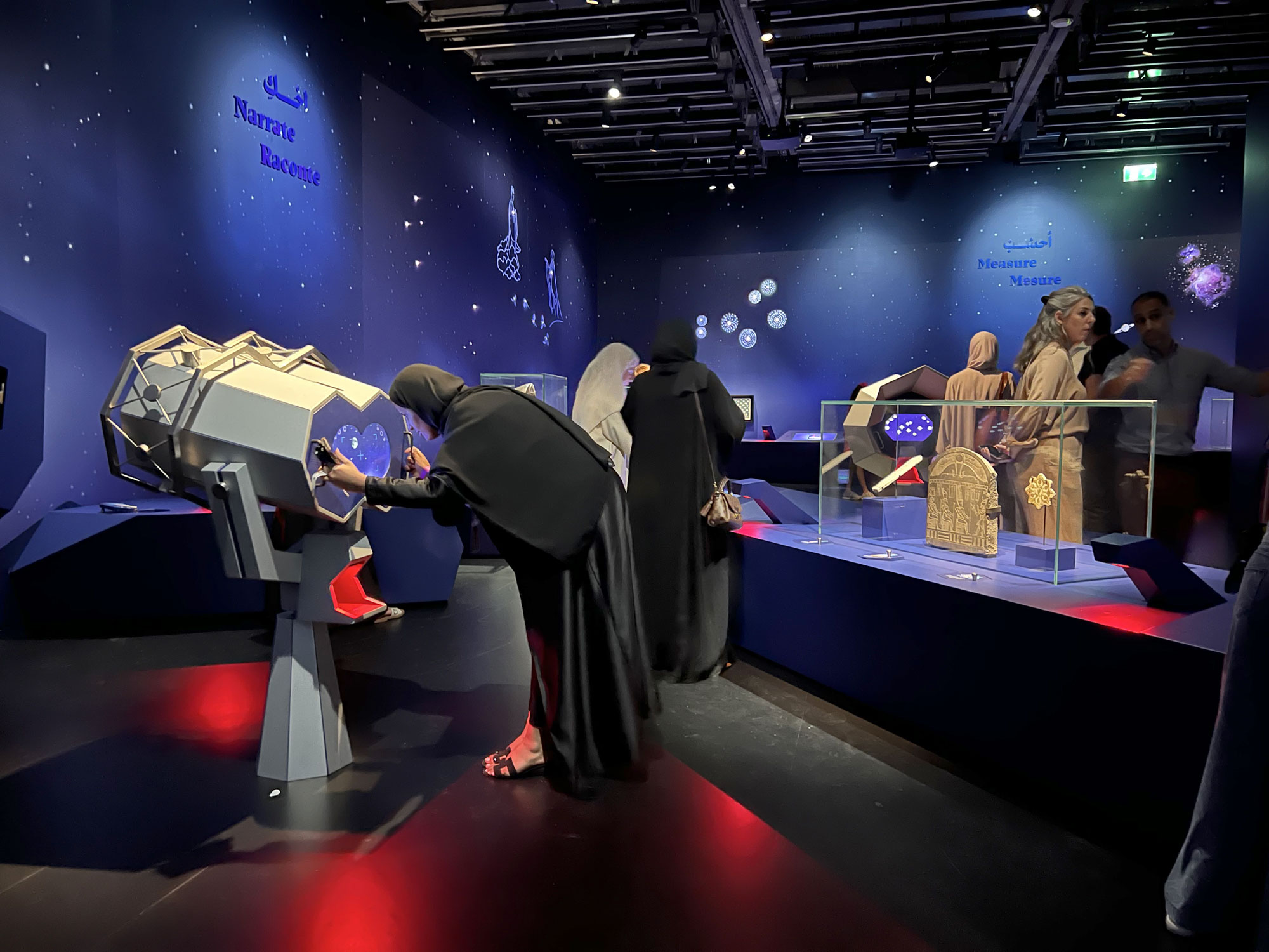 L'image montre une exposition dans un musée, probablement dédiée à l'astronomie ou à la science. Des visiteurs, dont plusieurs femmes portant des hijabs, interagissent avec des appareils de visualisation. En arrière-plan, on peut voir des objets exposés sous une lumière douce et une décoration étoilée sur les murs. L'ambiance semble à la fois éducative et attrayante, avec des présentations mettant en valeur des instruments scientifiques ou des artefacts culturels.