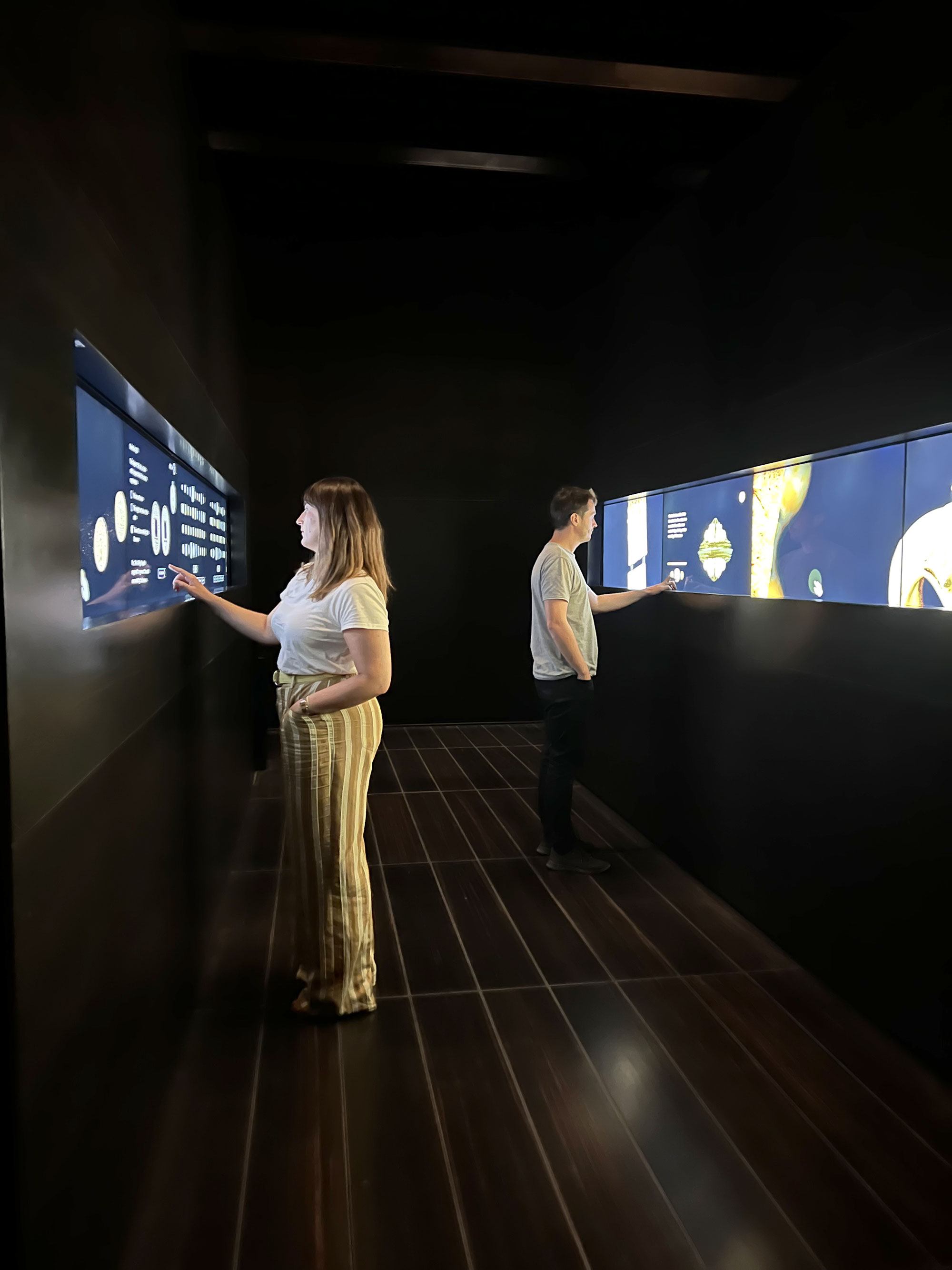 L'image montre deux personnes se tenant dans un espace intérieur sombre. Elles interagissent avec des écrans disposés sur les murs, qui affichent des informations visuelles. La femme, à gauche, porte un t-shirt blanc et un pantalon à rayures, tandis que l'homme, à droite, est vêtu d'un t-shirt gris et de jeans clairs. L'éclairage est subtil, créant une ambiance moderne et immersive.
