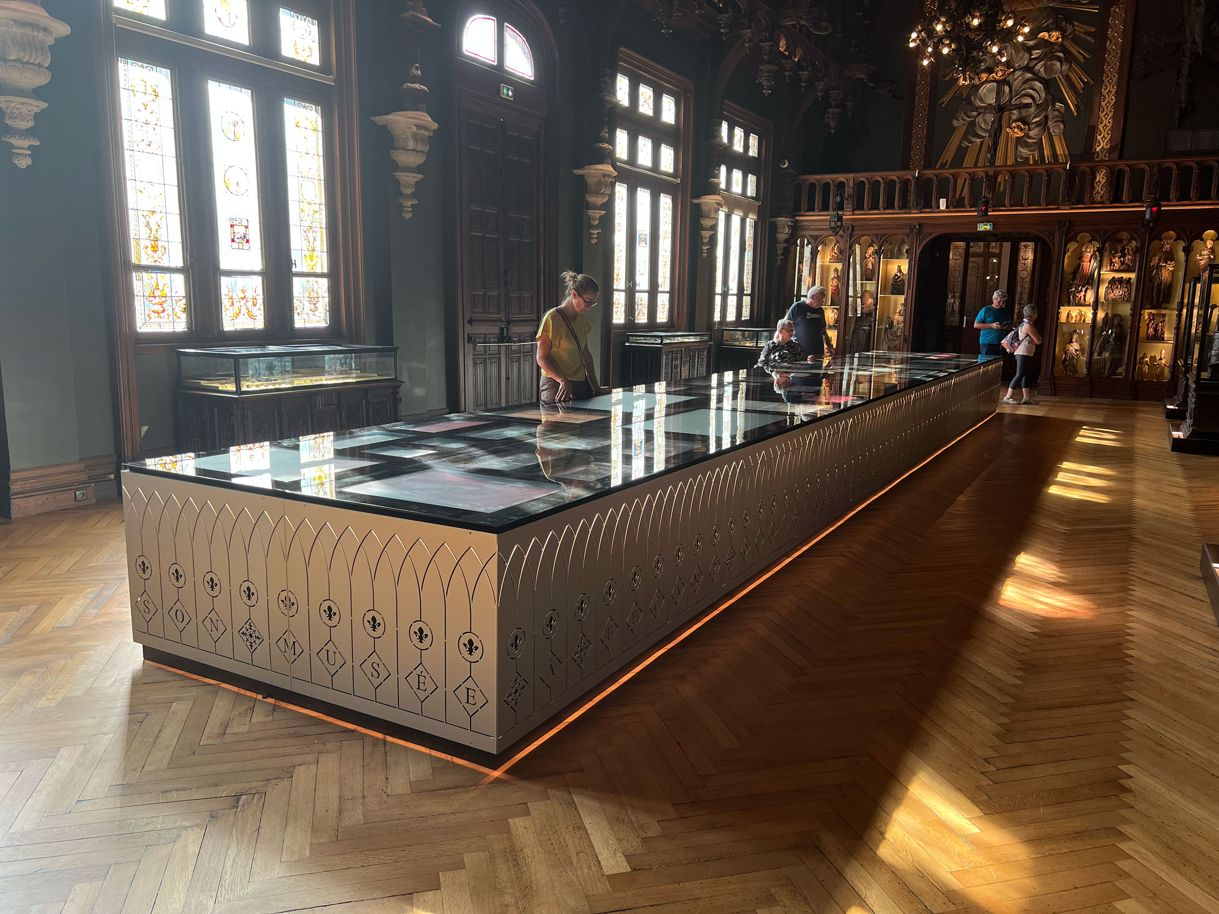 L'image montre une salle intérieure d'un musée ou d'un palais, avec un éclairage naturel provenant des fenêtres. Au centre, il y a une grande vitrine rectangulaire en verre qui semble exposer des objets, peut-être des artefacts ou des œuvres d'art. Le mobilier présente un design élégant avec des motifs décoratifs sur les côtés. Le sol est en bois avec un motif de parquet en chevron. Des visiteurs inspectent la vitrine, offrant une idée de l'ambiance paisible et muséale du lieu. Les murs sont ornés de détails architecturaux raffinés.