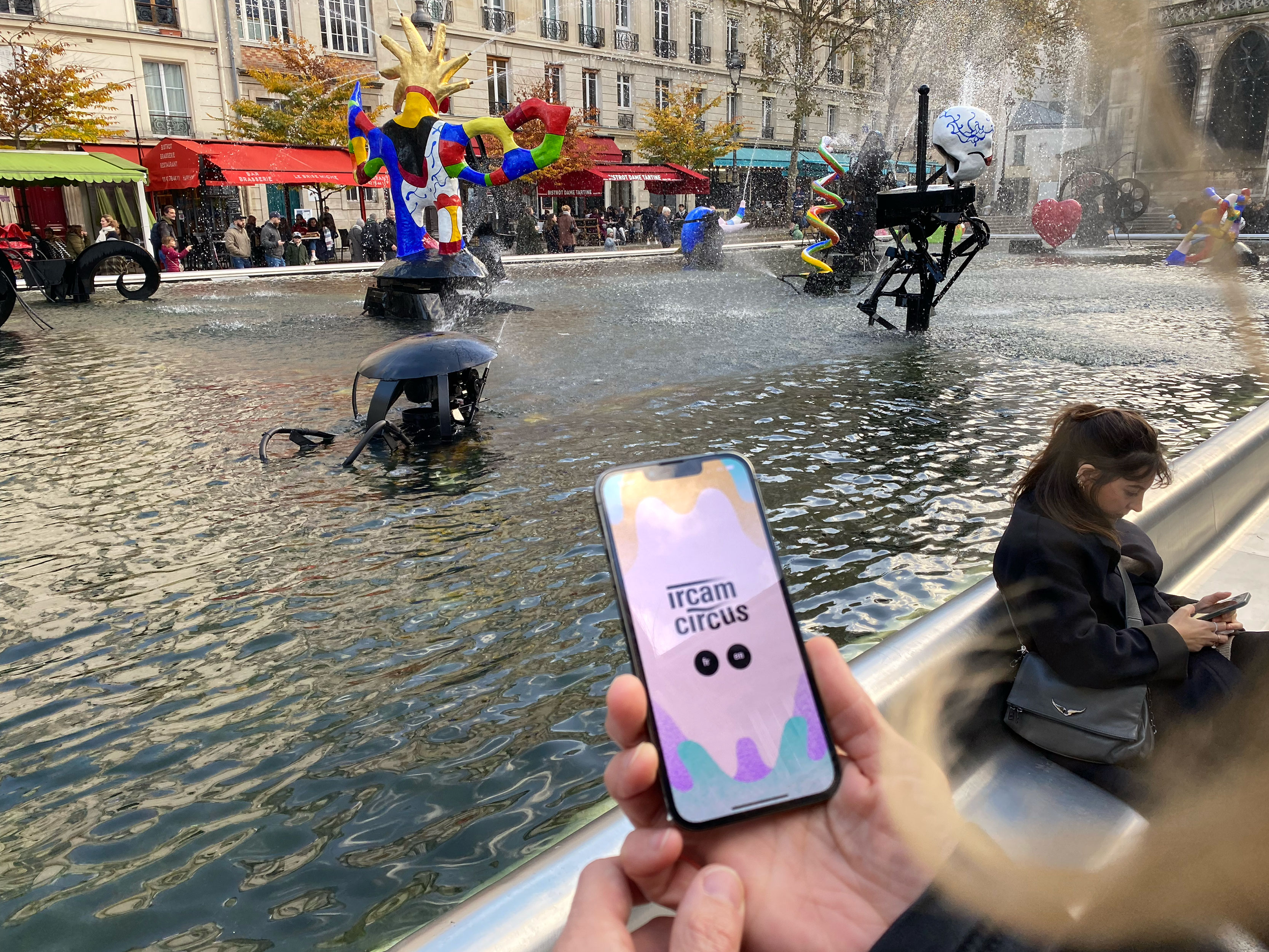 L'image montre une scène animée d'une place publique avec une fontaine au centre. On peut voir plusieurs sculptures colorées disposées autour de la fontaine. À l'avant-plan, une personne tient un smartphone affichant l'application "ircam circus". À droite, une autre personne est assise, probablement en train d'observer la scène. L'ambiance semble vivante et artistique, avec des couleurs vives et des éléments d'eau en mouvement.